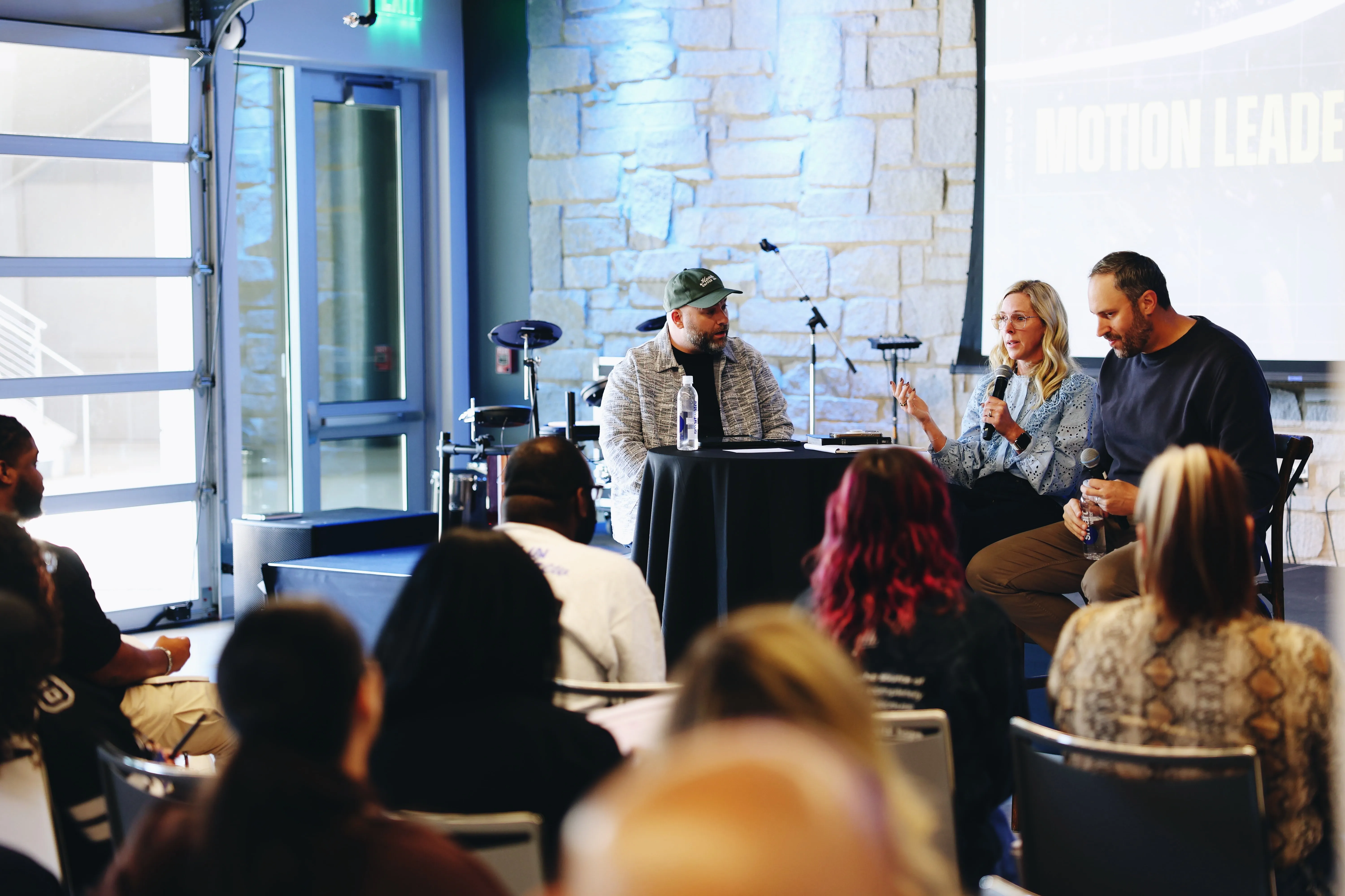 A panel discussion with three speakers seated at a round black table in front of an audience; one woman wearing glasses and a blue blouse holds a microphone and speaks, flanked by two men, one with a cap and a water bottle, the other holding a microphone and a bottle. The setting is indoors with a stone wall backdrop, a drum set, and a projected screen with partially visible text.