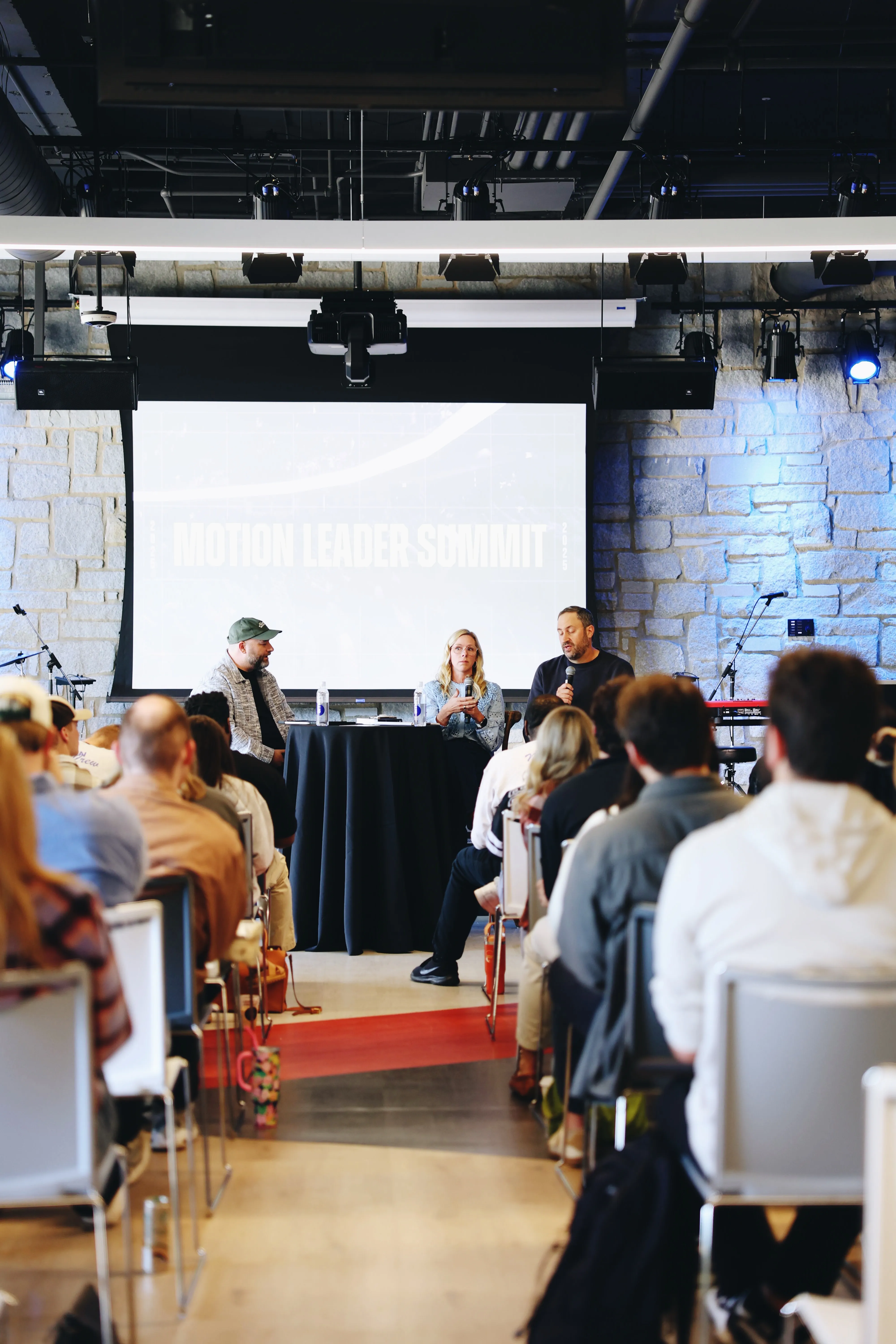 Three panelists sitting behind a tall round table with microphones in a conference room, speaking to an audience seated in rows facing them, with a large screen behind displaying 'Motion Leader Summit'.