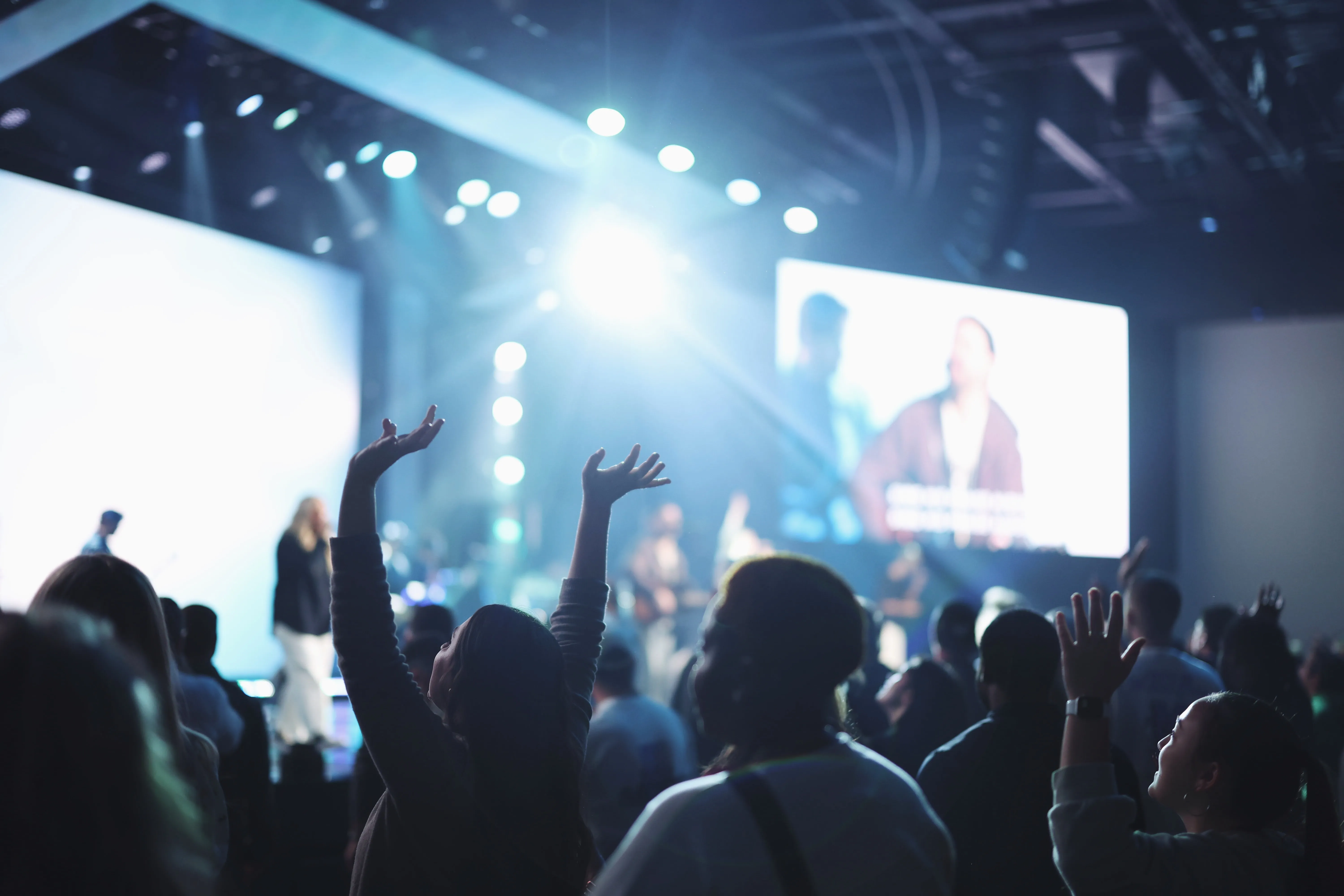 Audience at a concert or worship event raising their hands towards a brightly lit stage with performers and a large screen in the background.