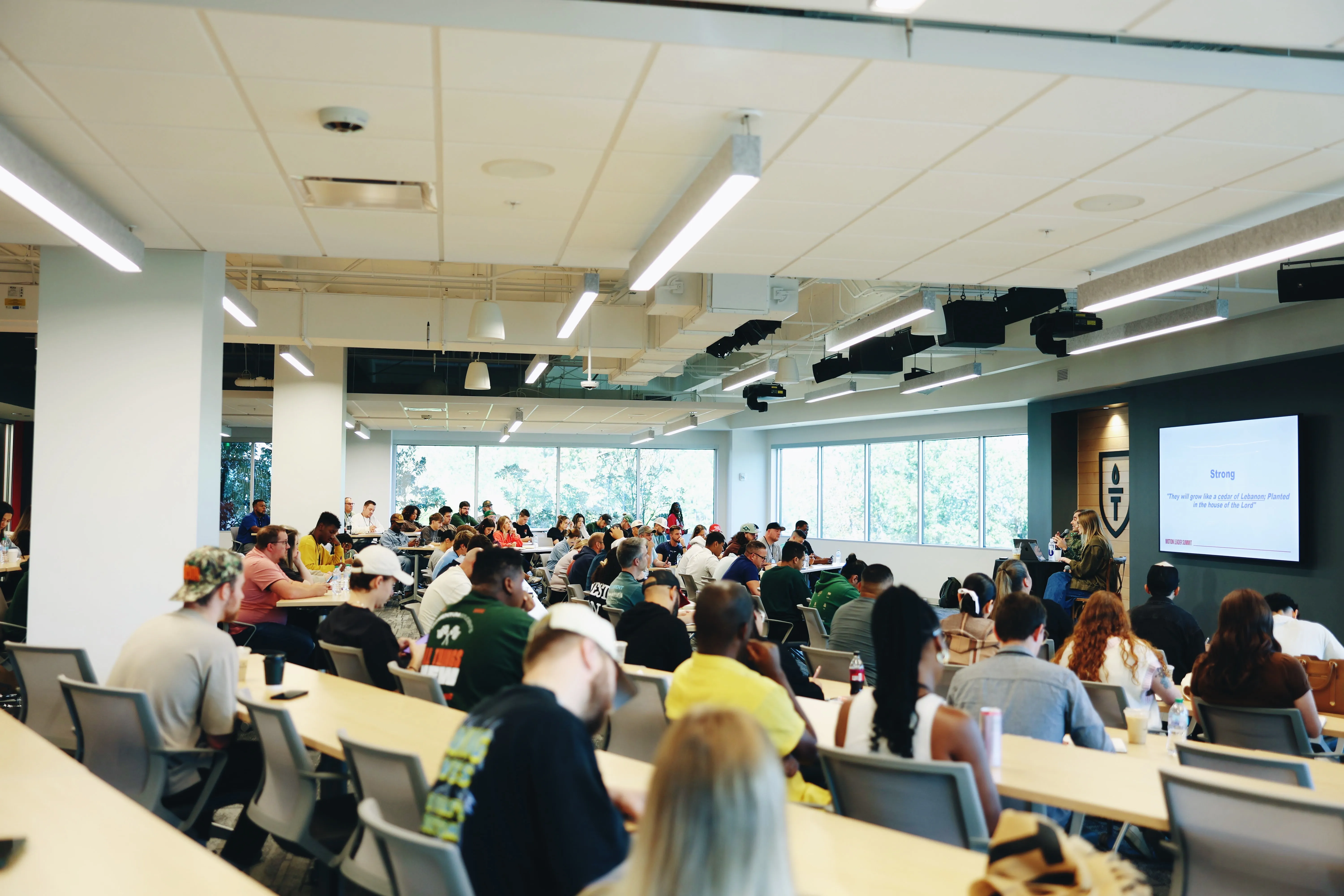 Wide classroom filled with students sitting at long tables facing the front where a presenter talks next to a screen displaying a slide titled 'Strong' with a quote about growing like a cedar of Lebanon.
