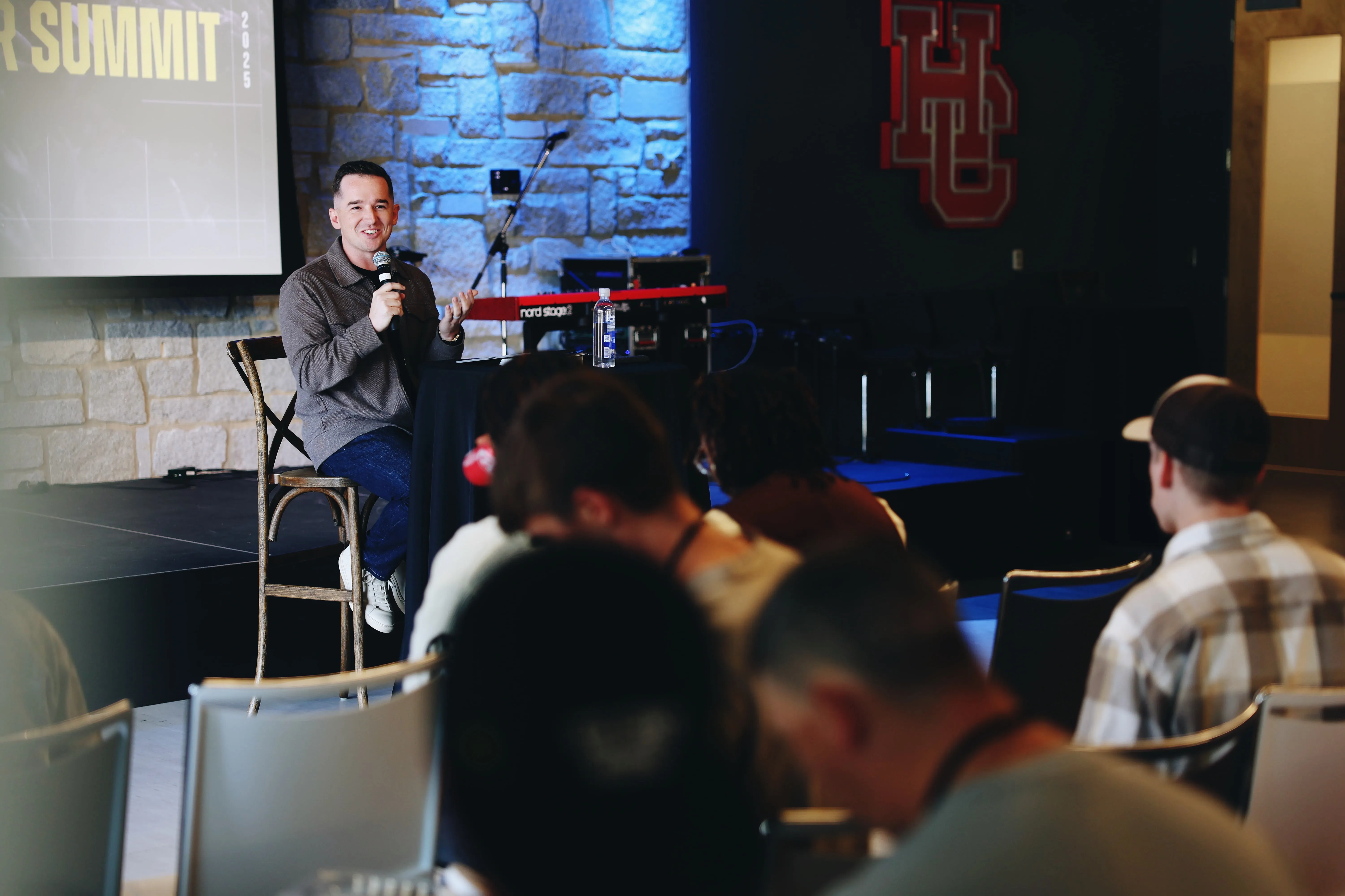 Man sitting on a wooden chair on stage speaking into a microphone during a presentation, with audience seated facing him, a water bottle on a small round table, a keyboard in the background, and partially visible text reading 'SUMMIT' on a screen.