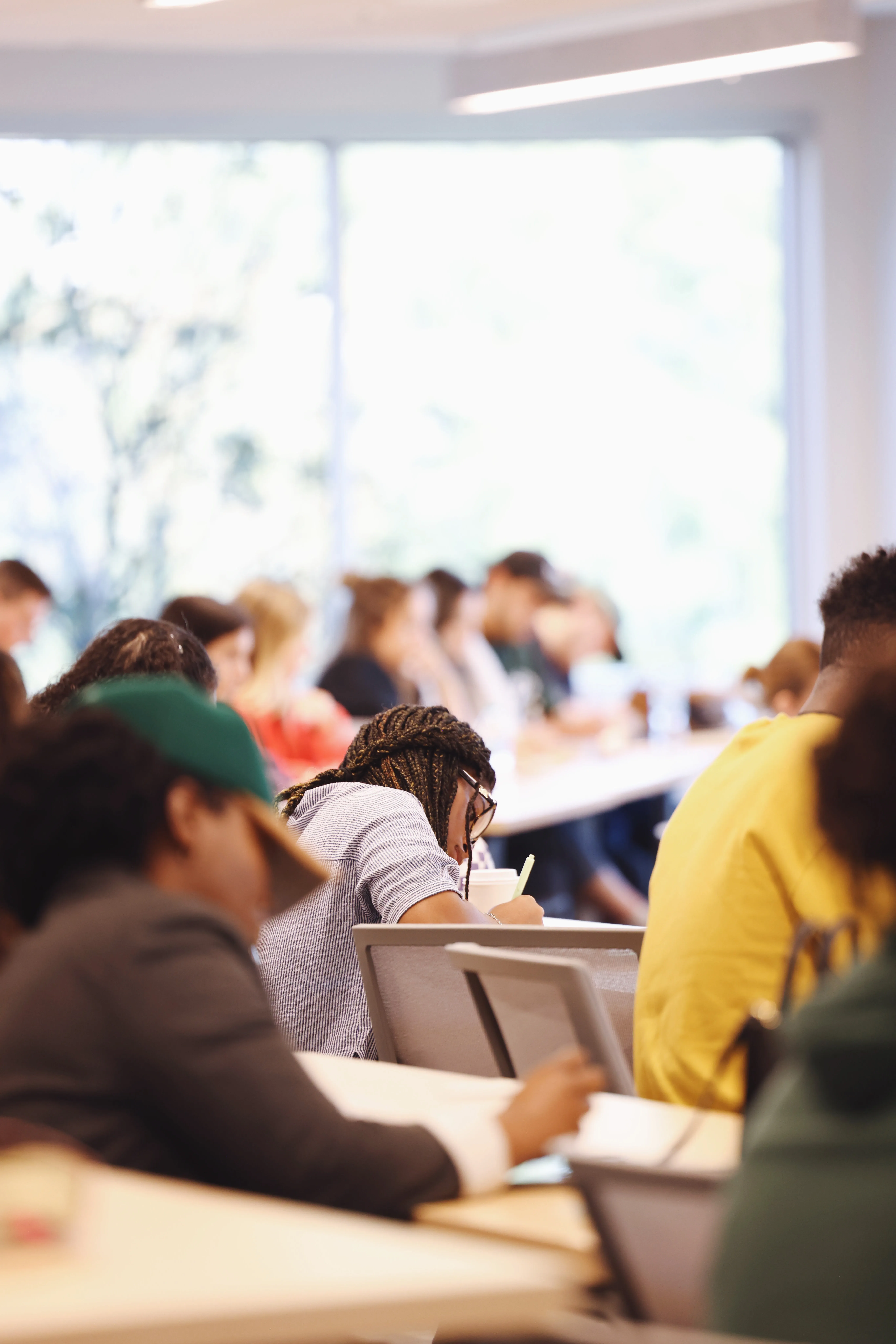 Students seated in a classroom, focusing on writing or taking notes, with a large window in the background letting in natural light.