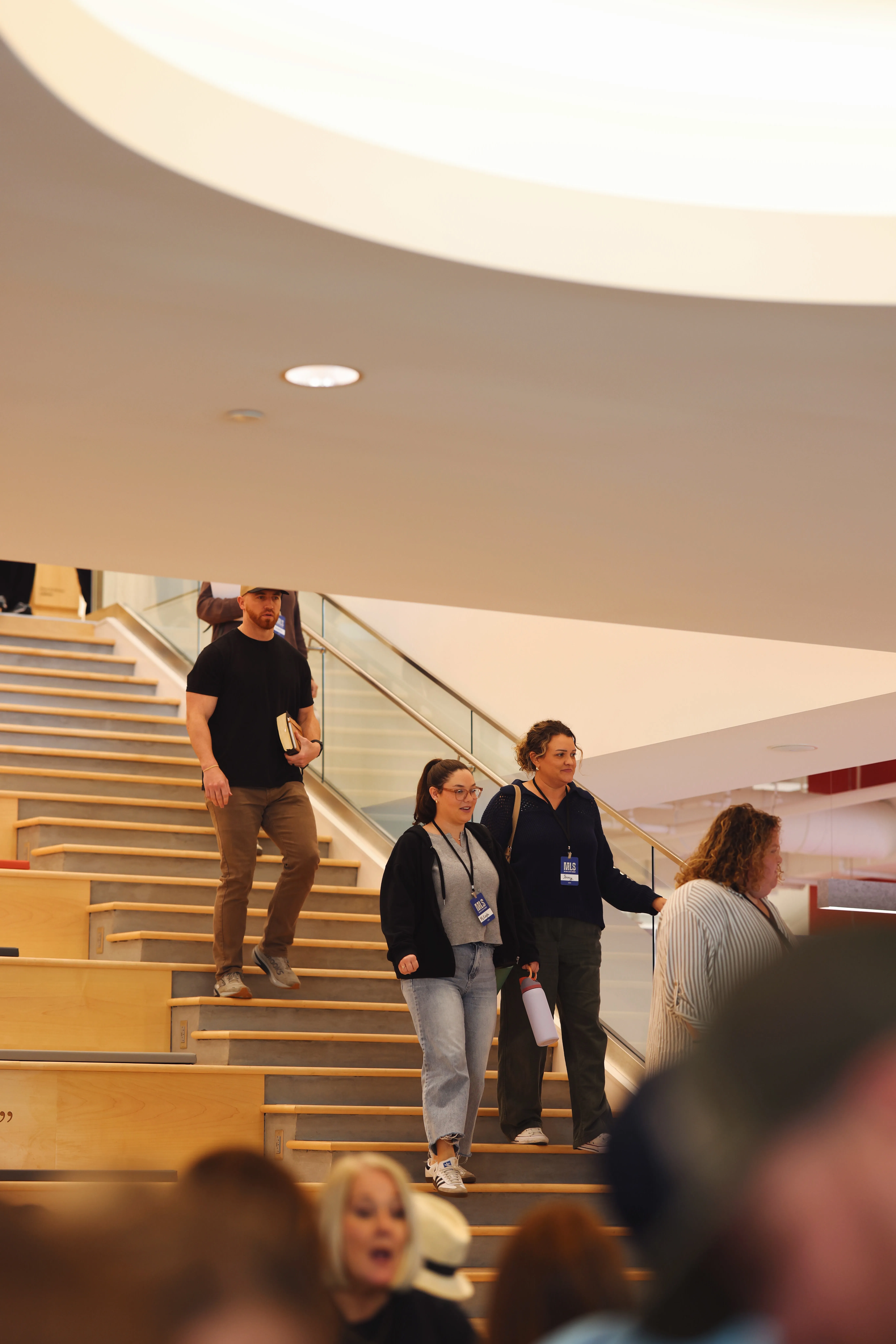 Three people walking down indoor wide stairs with wooden steps; two women walk side by side in front, one holding a water bottle, and a man follows behind holding a book, all wearing visitor badges on lanyards.