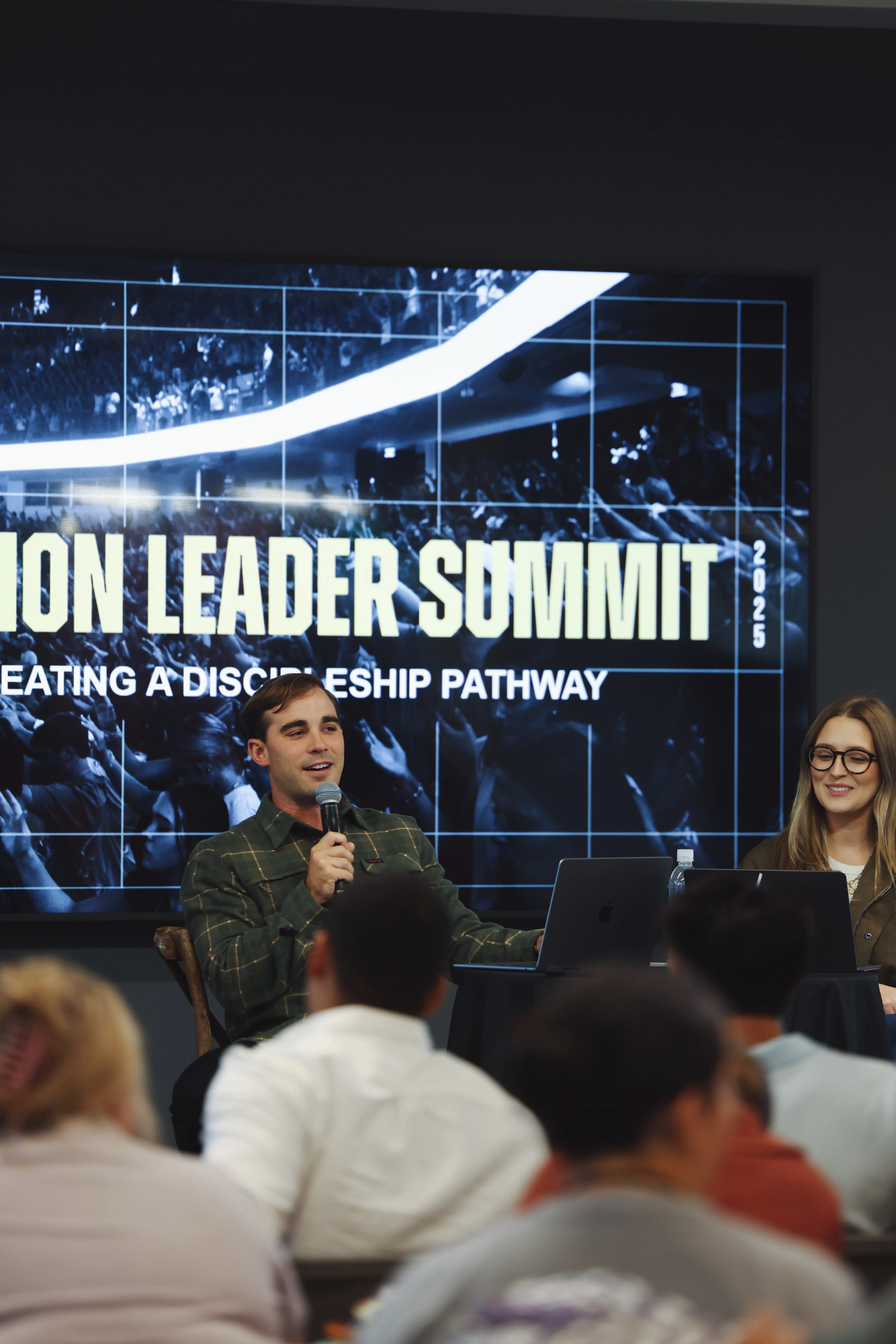 A man holding a microphone and speaking at a leadership summit with a woman sitting next to him, both facing an audience with laptops on the table, and a large screen behind them displaying the words 'LEADER SUMMIT' and 'CREATING A DISCIPLESHIP PATHWAY 2025'.
