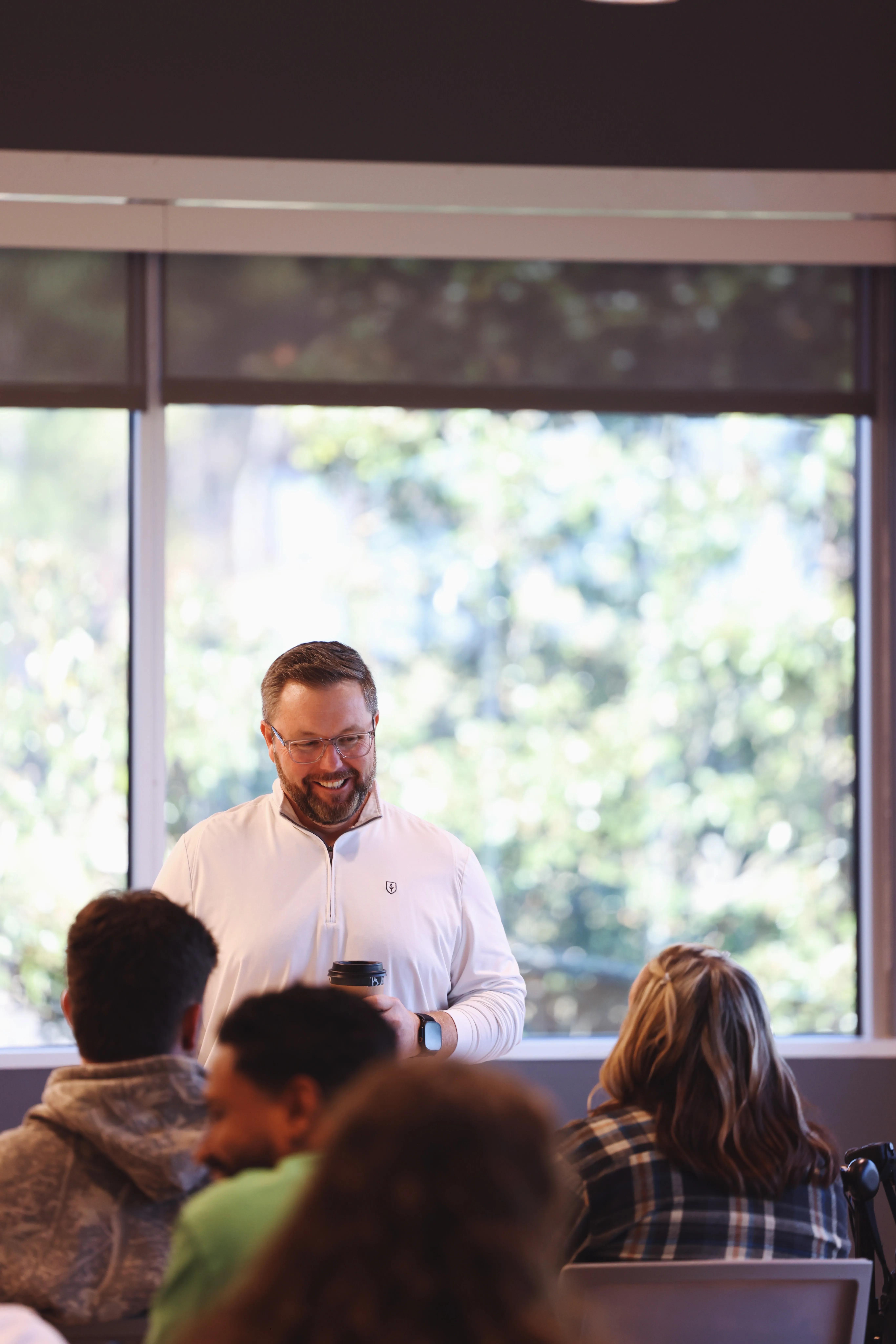 Man with glasses and a beard smiling and holding a coffee cup in a room with large windows, facing a group of seated people.