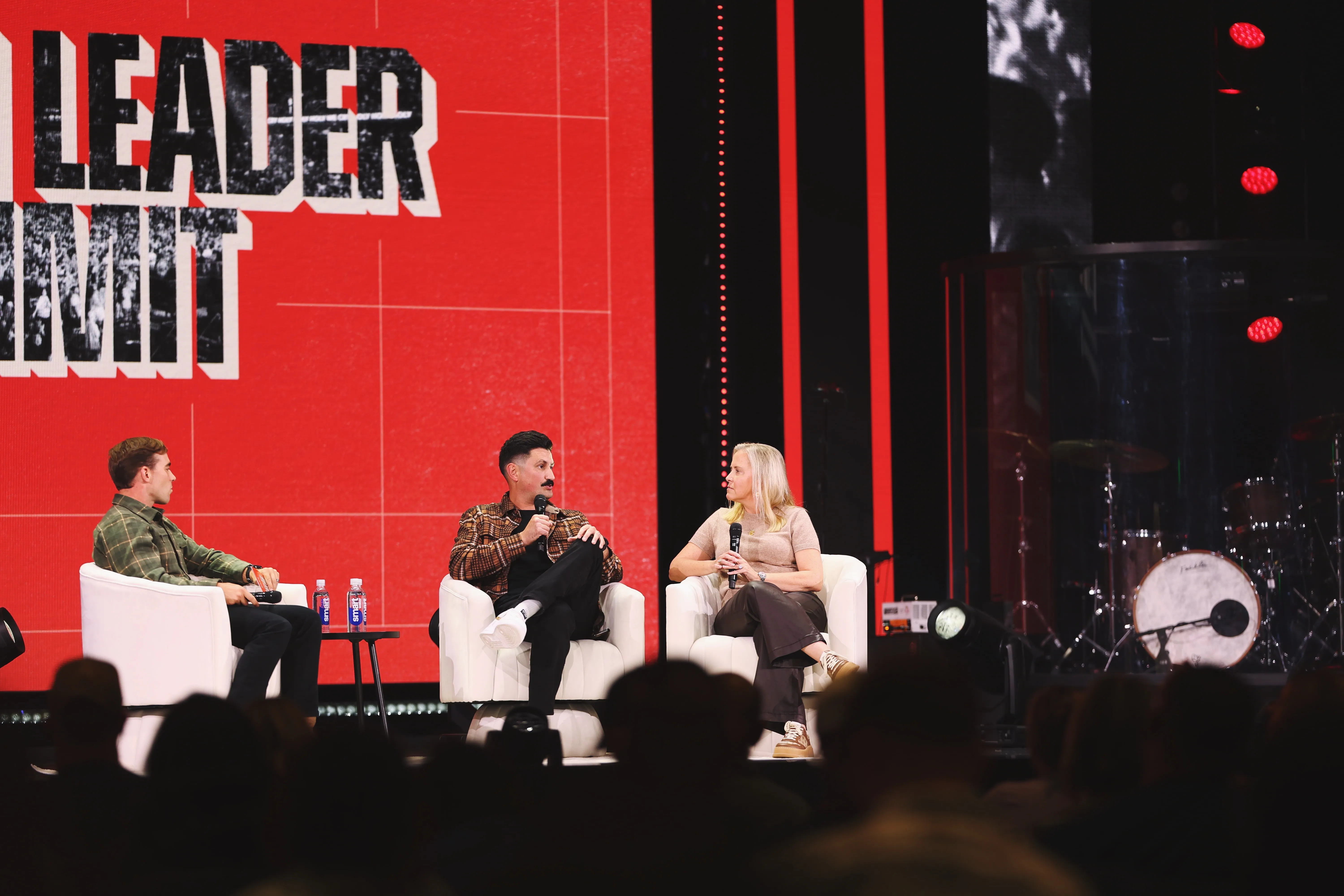 Three people seated on white armchairs on a stage with a large red backdrop displaying the partial text 'LEADER SUMMIT', two men and one woman, each holding microphones, engaged in a discussion with a visible drum set in the background and an audience silhouette in the foreground.