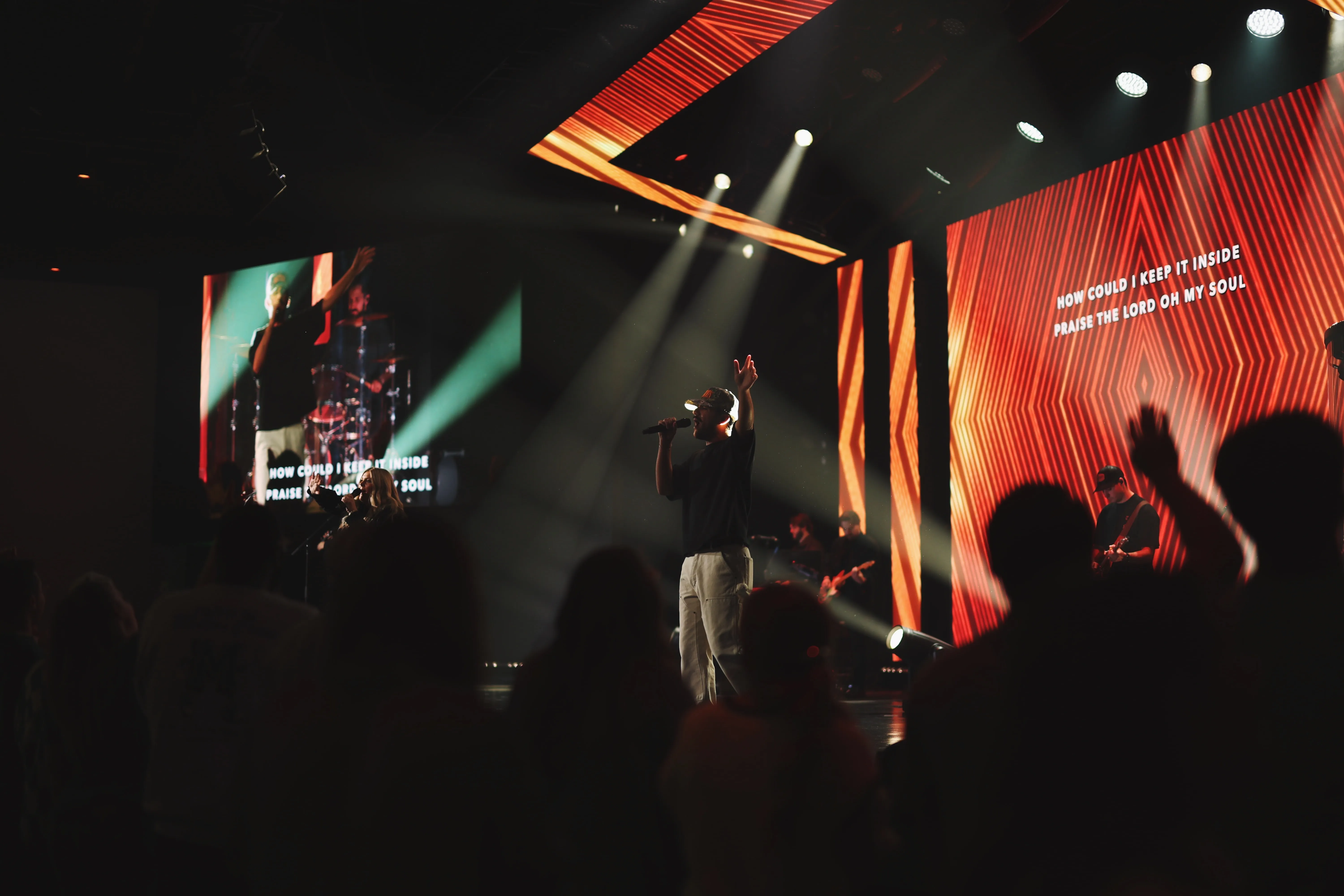 Musician wearing a cap and holding a microphone with one hand raised performs on stage with a band, illuminated by spotlights, while abstract red and orange patterns and song lyrics are displayed on large screens behind him, in front of a dark audience.