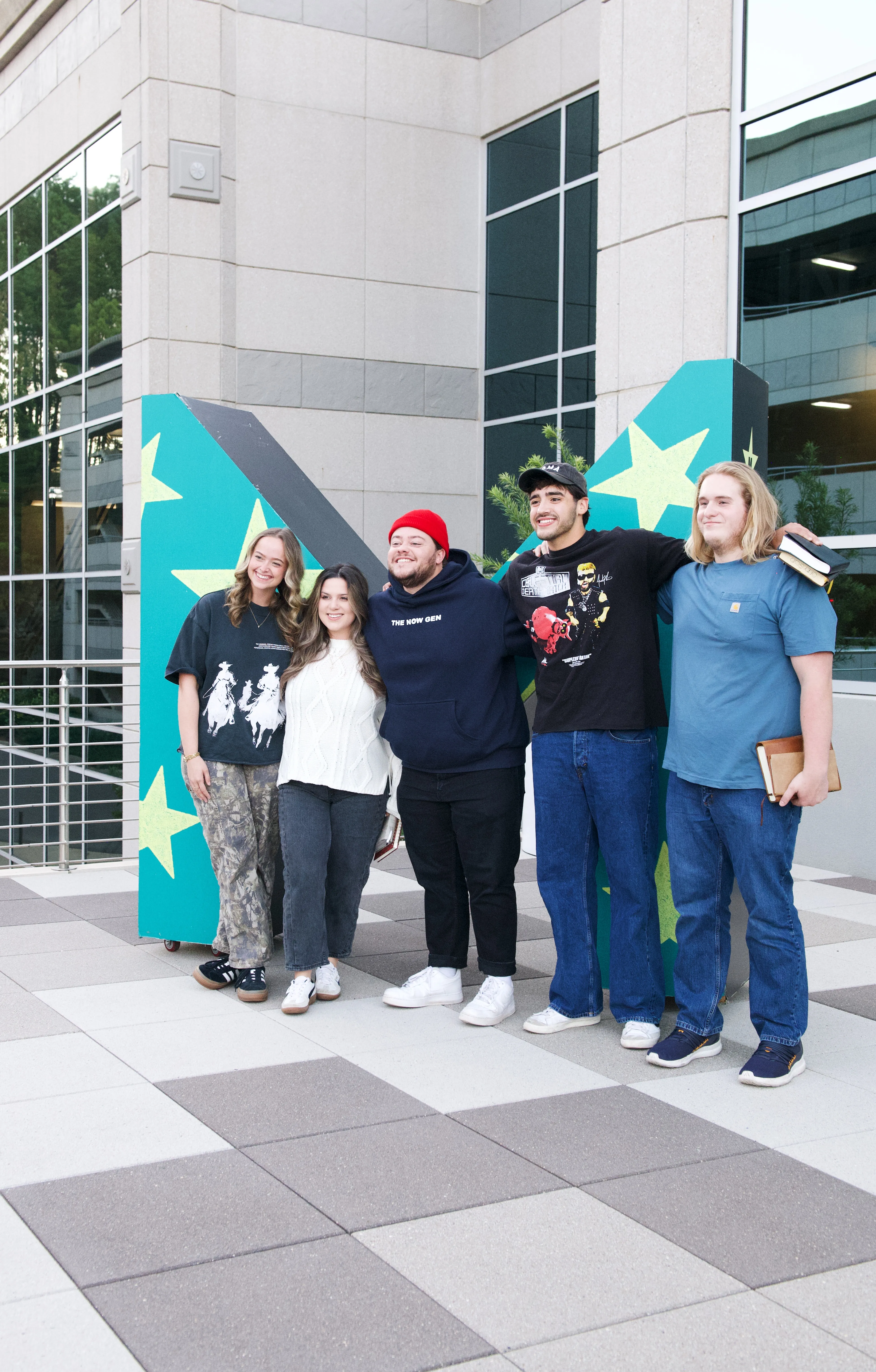 Five young adults standing side by side outdoors in front of a large painted letter with green and yellow star designs, smiling and posing for the photo; two hold books, and the group is dressed casually.