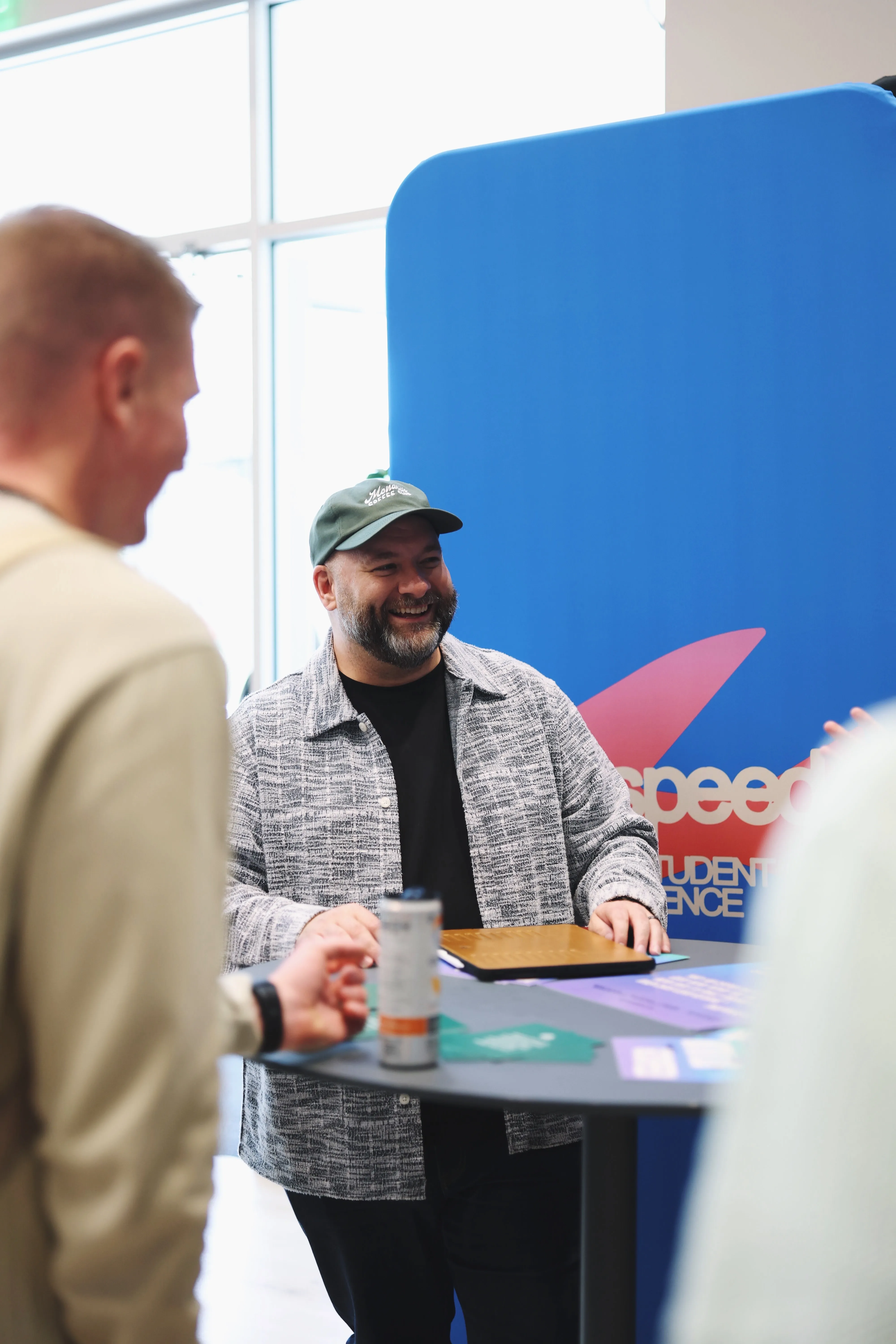 A man with a beard, wearing a green cap and a gray patterned jacket, smiling and standing at a high table with a closed laptop and drink can, talking to two other people partially visible. A blue background with red and white text is behind him.
