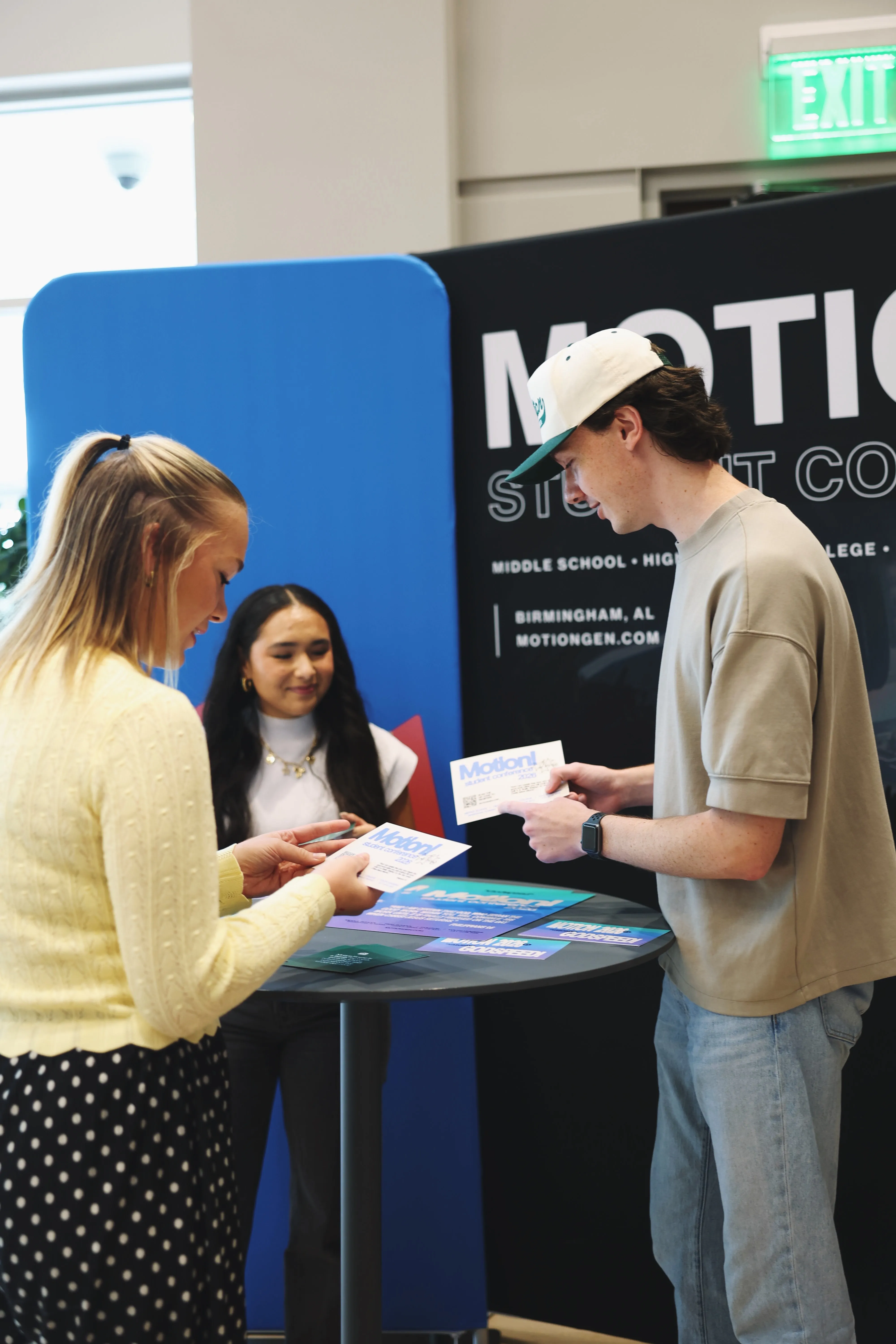 Three young people standing around a tall round table, holding and looking at flyers for Motion Student Conference, with a backdrop showing the event name and location in Birmingham, AL.