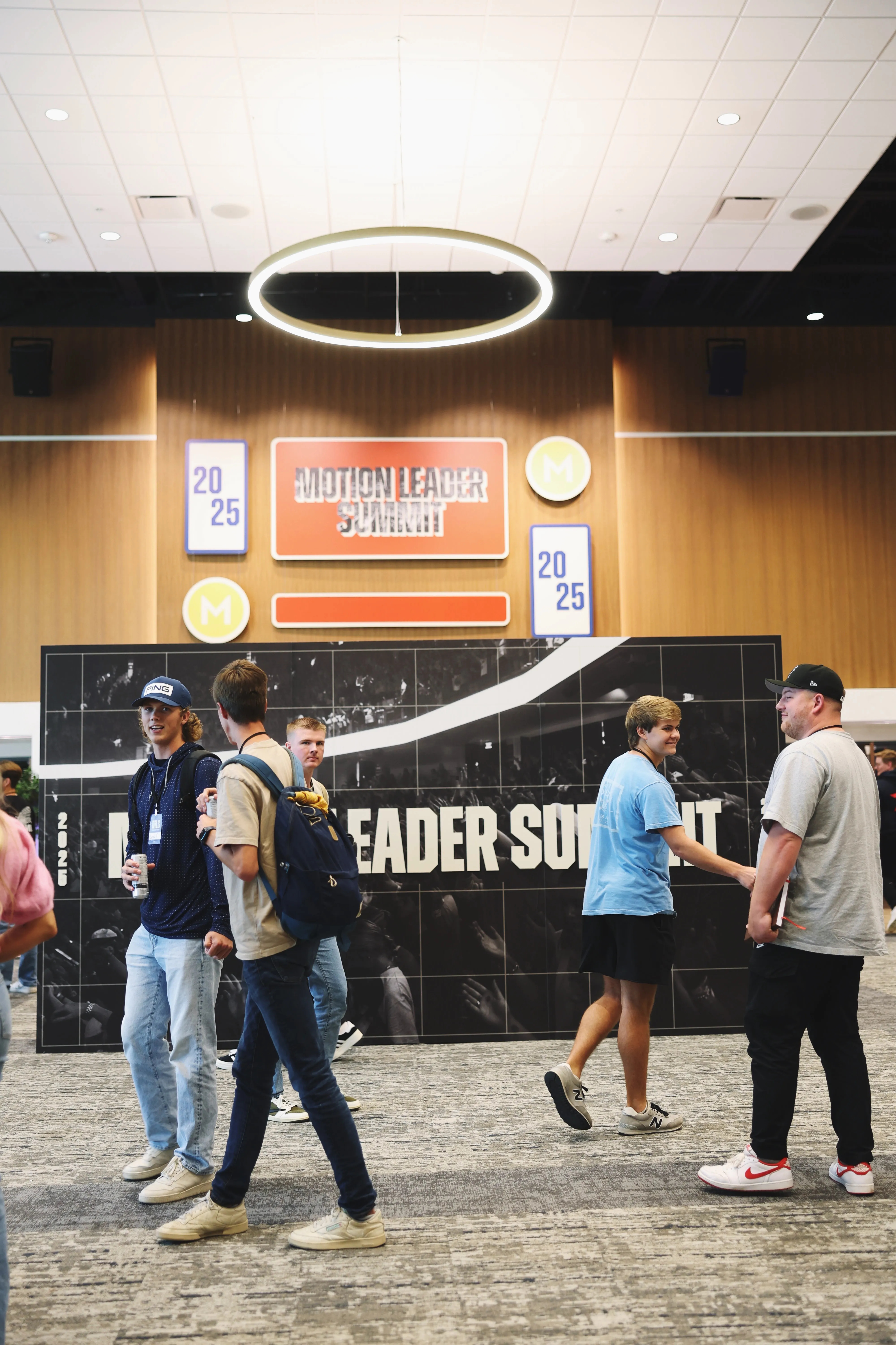 Group of young people talking and walking inside a conference venue with a large sign reading 'Motion Leader Summit' and the year 2025 displayed on the wall behind them.