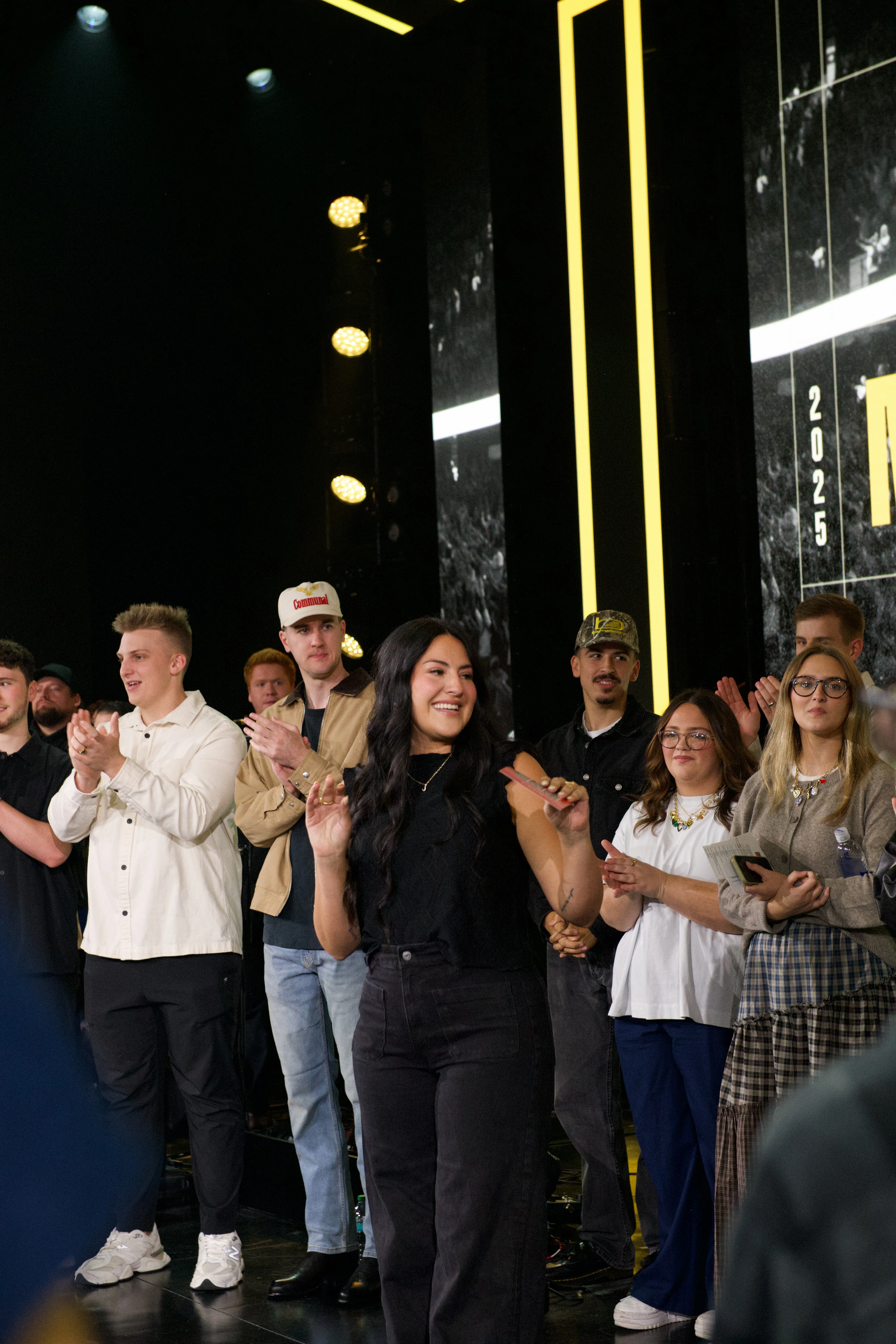 Group of people standing on a stage with black and yellow background, clapping and smiling, including a woman in black pants and top holding a phone.