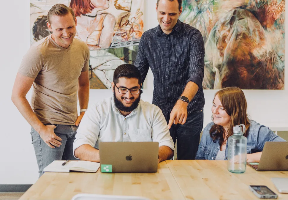 4 people standing and sitting near the table and looking at a laptop
