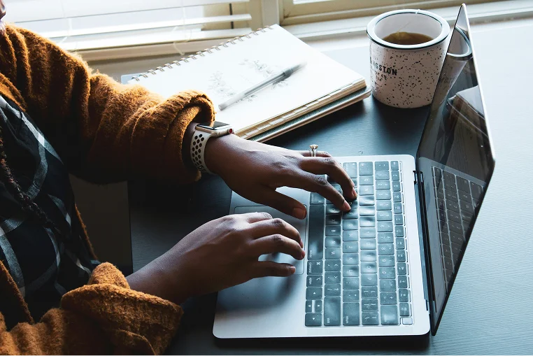 Close-up of hands typing on a laptop keyboard, with a coffee cup and papers on the desk nearby