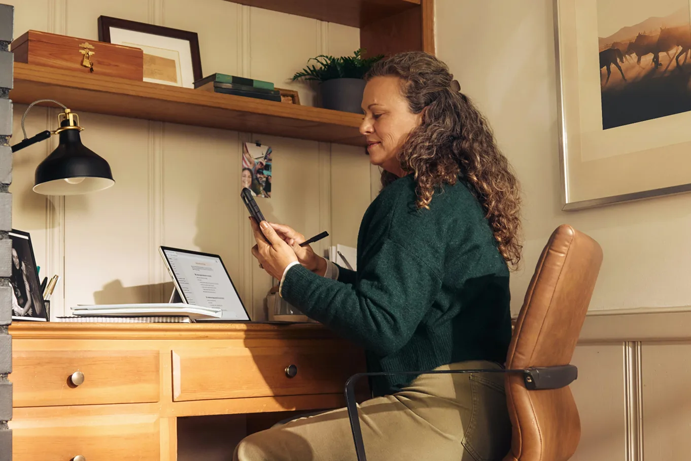 A woman with curly hair sits at a wooden desk in a home office, looking at her smartphone. A laptop is open on the desk, with a lamp, framed photo, and shelf with books visible in the background.