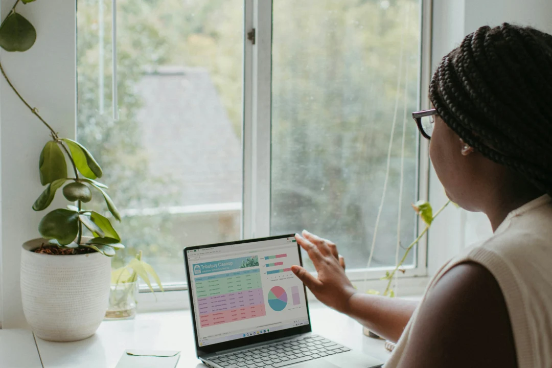 A woman reviews a data dashboard on her laptop at a bright, plant-filled desk.