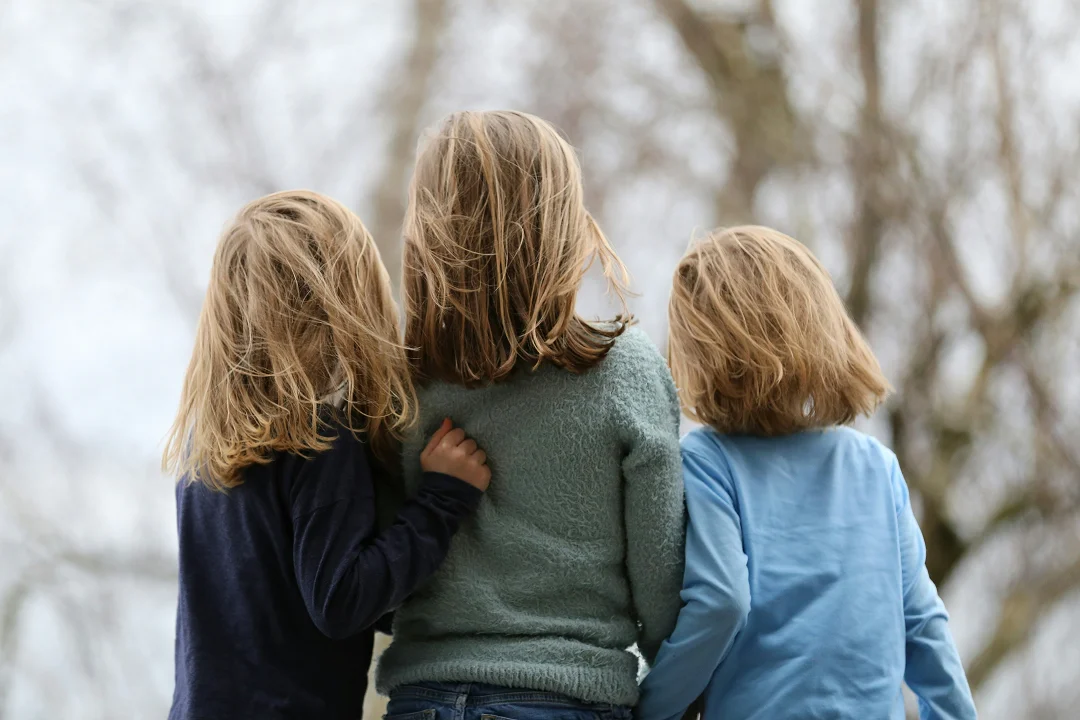 Three children stand together outdoors, facing away from the camera with their arms around each other.Three children stand together outdoors, facing away from the camera with their arms around each other.