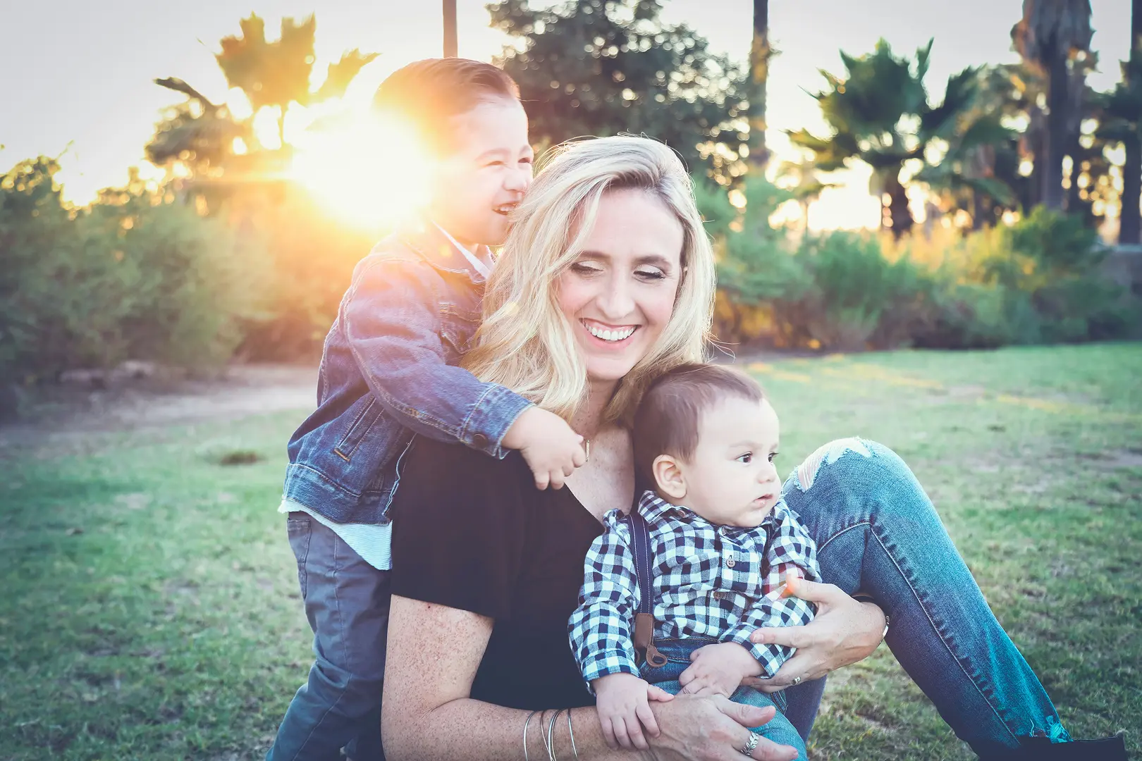A mother sits on grass in a sunny park, smiling as her young son hugs her from behind while she holds her baby on her lap.