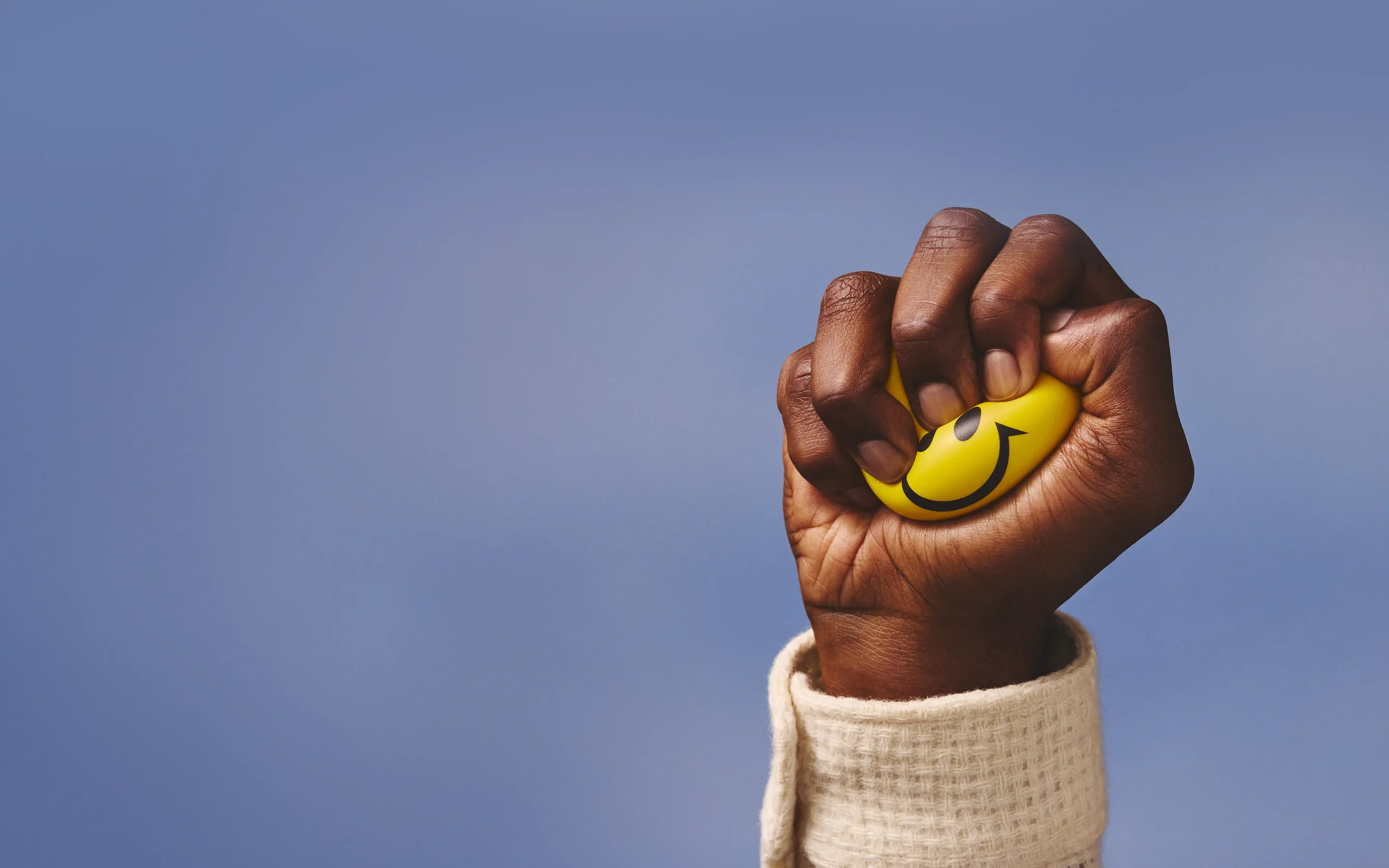 A hand squeezing a smiley-face stress ball against a soft blue background