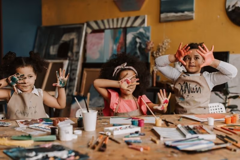 Three children sitting at a table with art supplies, showing their hands covered in colorful paint and smiling.