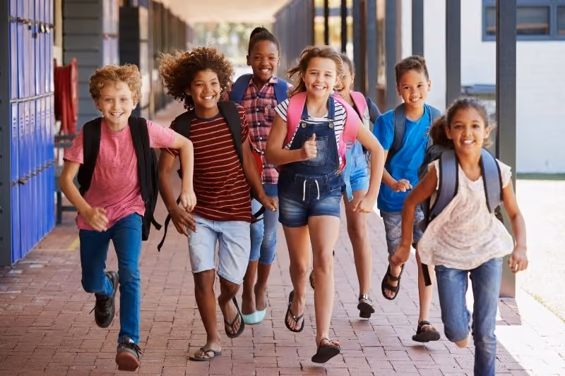 Six happy children running down a school hallway with backpacks, smiling and holding hands.