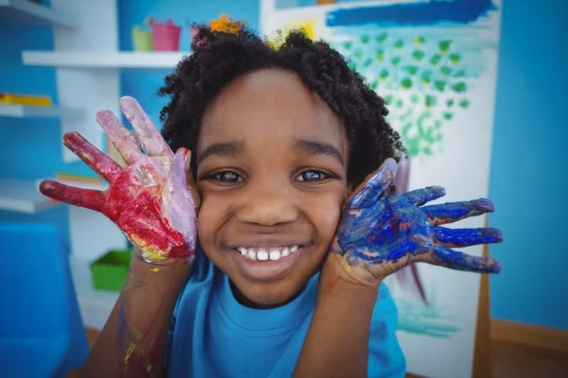 Smiling young child with curly hair showing hands covered in red, purple, and blue paint in a colorful room.