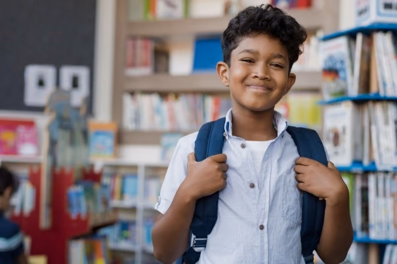 Smiling boy holding straps of his dark blue backpack in a library filled with bookshelves.