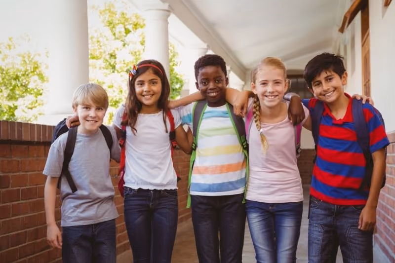 Five smiling schoolchildren standing close together in a school corridor with arms around each other's shoulders, carrying backpacks and dressed casually, with a row of columns and windows in the background.