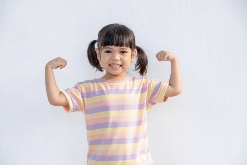Smiling young girl with pigtails wearing a pastel striped t-shirt flexing her arm muscles against a plain light background.