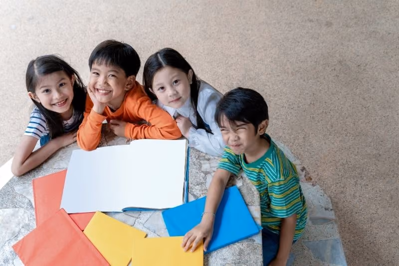 Four young children sitting around a round stone table with colorful folders and a large open blank book, smiling and looking up at the camera.