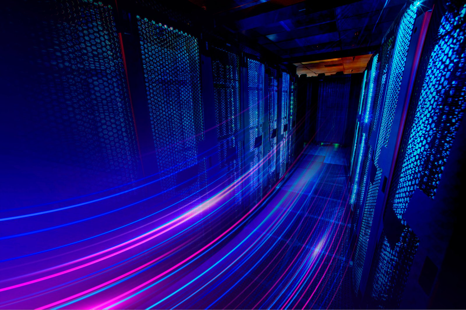 A imagem mostra o corredor interno de um data center ou sala de servidores. Há fileiras simétricas de racks de servidores dos dois lados, formando um corredor estreito que leva até uma porta ao fundo. Os equipamentos possuem várias luzes de LED azuis e vermelhas piscando, indicando atividade e funcionamento constante. A iluminação é fria, predominantemente azul, vinda de painéis retangulares no teto, o que cria um ambiente tecnológico e moderno. O piso é elevado, com placas modulares típicas de data centers (provavelmente para passagem de cabos e ventilação), e há uma grade de ventilação no chão no primeiro plano. A porta no fundo também tem um painel iluminado com linhas e pontos que lembram circuitos eletrônicos, reforçando a estética digital e futurista da cena. No geral, a imagem transmite: alta tecnologia segurança e controle infraestrutura de armazenamento/processamento de dados clima frio e silencioso típico de ambientes de TI Parece um espaço dedicado à hospedagem de servidores, cloud ou infraestrutura de rede.