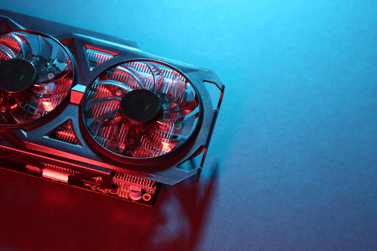 A imagem apresenta um close-up de uma placa de vídeo (GPU) posicionada sobre uma superfície lisa, destacando seu sistema de refrigeração composto por duas grandes ventoinhas transparentes. O enquadramento evidencia os detalhes do acabamento da carcaça, das aletas do dissipador de calor e de parte da placa de circuito impresso (PCB), transmitindo uma sensação de robustez e alto desempenho. A iluminação em tons de vermelho e azul cria um forte contraste visual e confere um aspecto futurista à cena, enquanto o amplo espaço negativo à direita torna a composição ideal para aplicações em banners, páginas de produtos ou materiais institucionais. A imagem remete a conceitos como computação de alta performance, inteligência artificial, aceleração por GPU, processamento gráfico, infraestrutura de TI, data centers e inovação tecnológica.