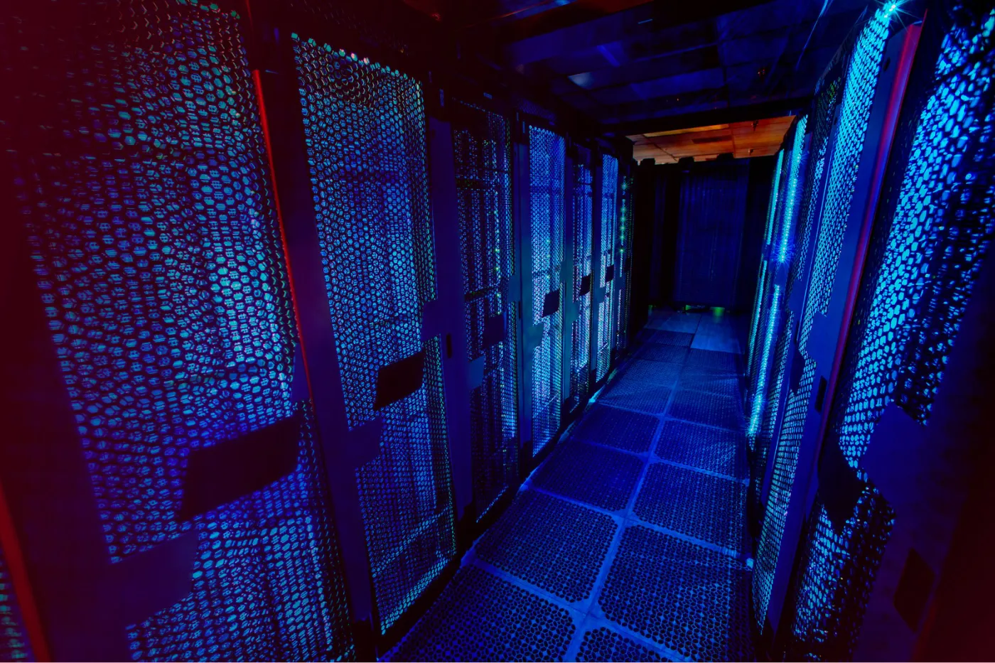 A imagem mostra o corredor interno de um data center. De ambos os lados, há fileiras de racks de servidores altos, protegidos por portas metálicas perfuradas em formato de malha hexagonal. A iluminação predominante é azul intensa, misturada com tons de vermelho nas laterais, criando um contraste tecnológico e futurista. As luzes dos equipamentos refletem nas superfícies metálicas, sugerindo atividade constante dos servidores. O piso é elevado, composto por placas modulares perfuradas, típico de ambientes de data center para circulação de ar e refrigeração. Ao fundo, o corredor se estreita até uma cortina ou divisória escura. A cena transmite sensação de infraestrutura robusta, alta performance e ambiente crítico de TI — associada a armazenamento de dados, computação em nuvem e operações de missão crítica.