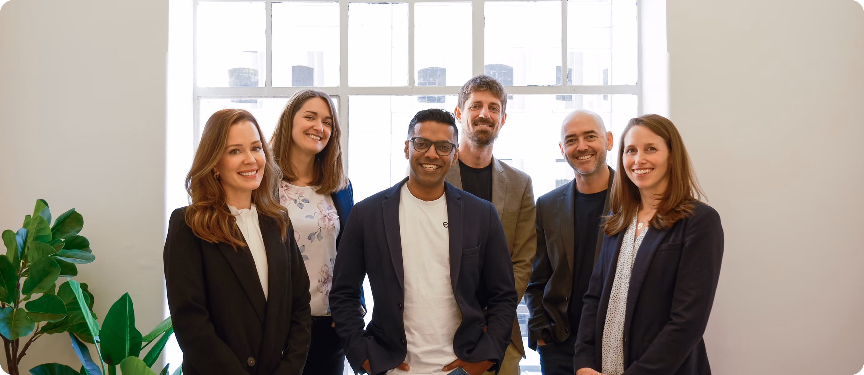 Six diverse professionals smiling and standing indoors with a large window and plant beside them.