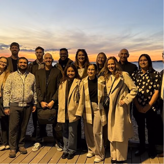 Group of people standing on a wooden dock at sunset by the water, smiling and posing for a photo.