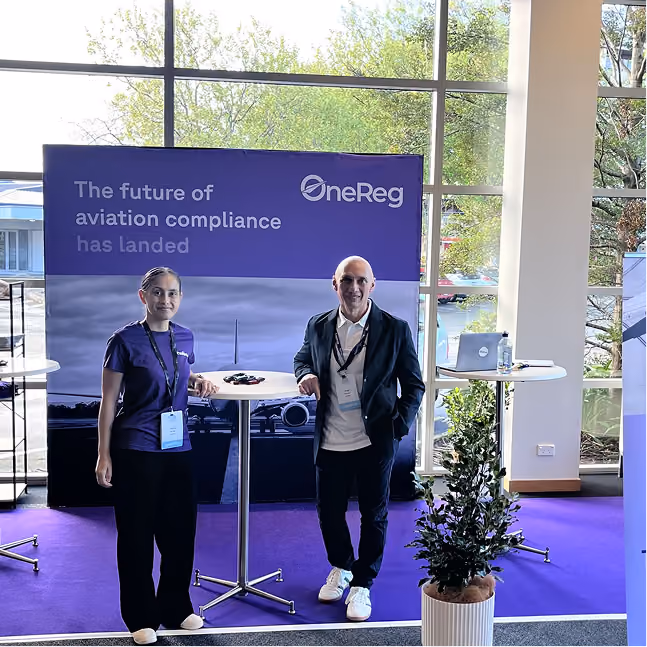 Two people standing by a table at a booth with an aviation compliance banner in the background.
