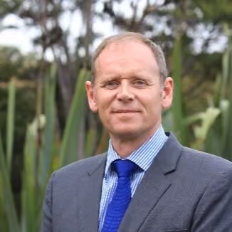 Man in a blue suit and tie standing outdoors with greenery and trees in the background.