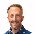 Smiling man with short blond hair and beard wearing a blue patterned shirt on a white background.
