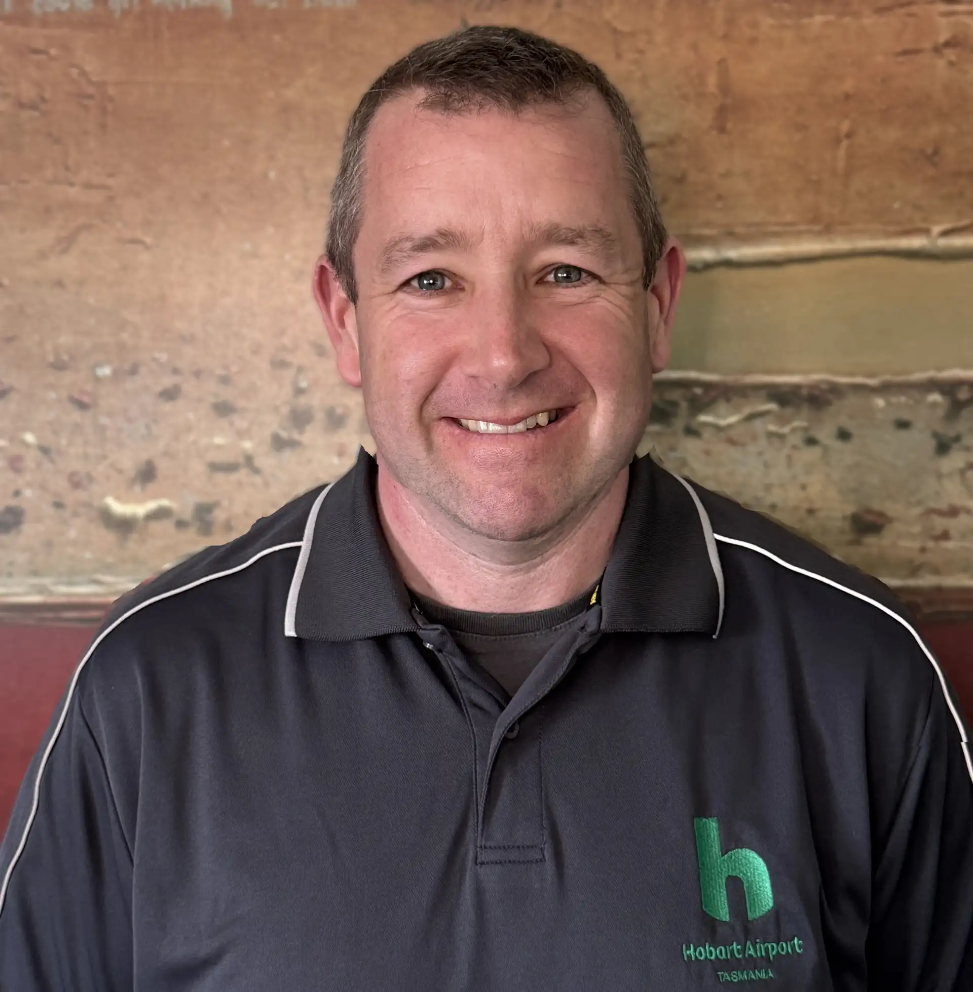 Smiling man wearing a Hobart Airport Tasmania polo shirt against a rustic wall background.