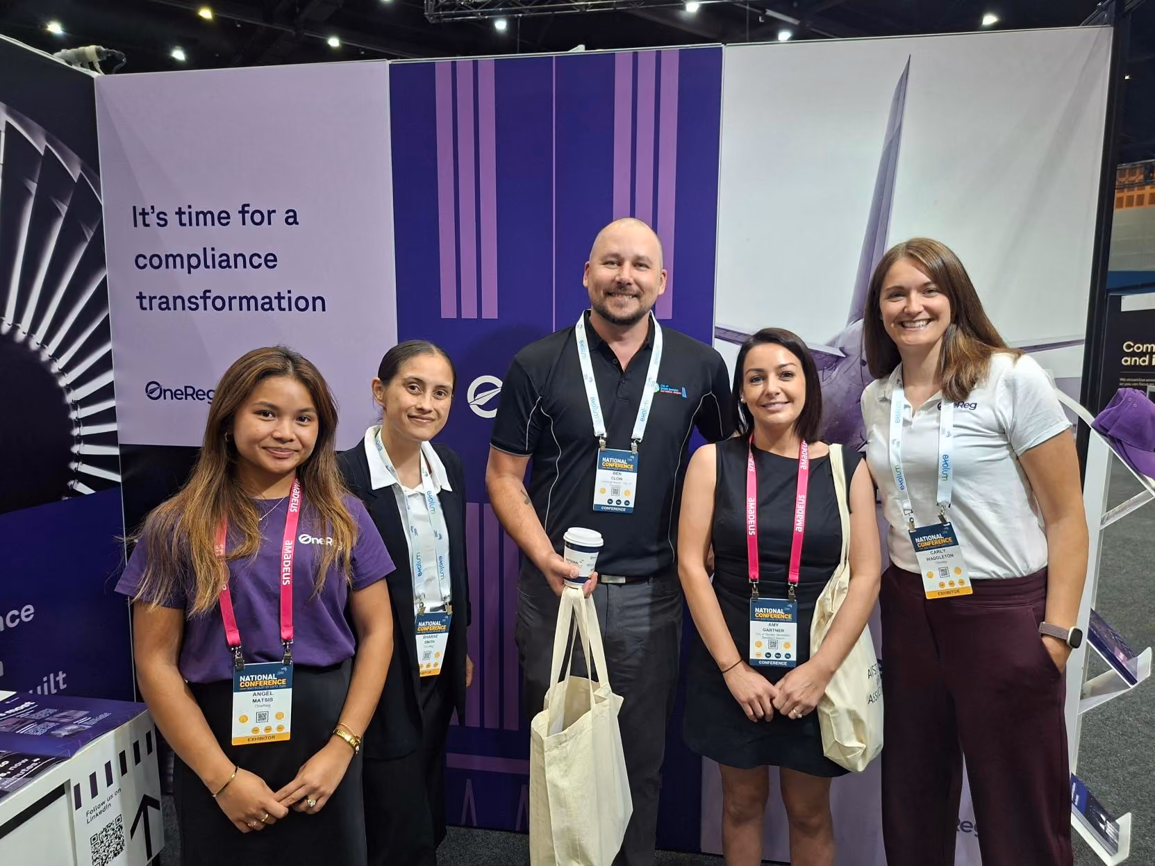 Five conference attendees smiling, standing in front of a banner about compliance transformation.