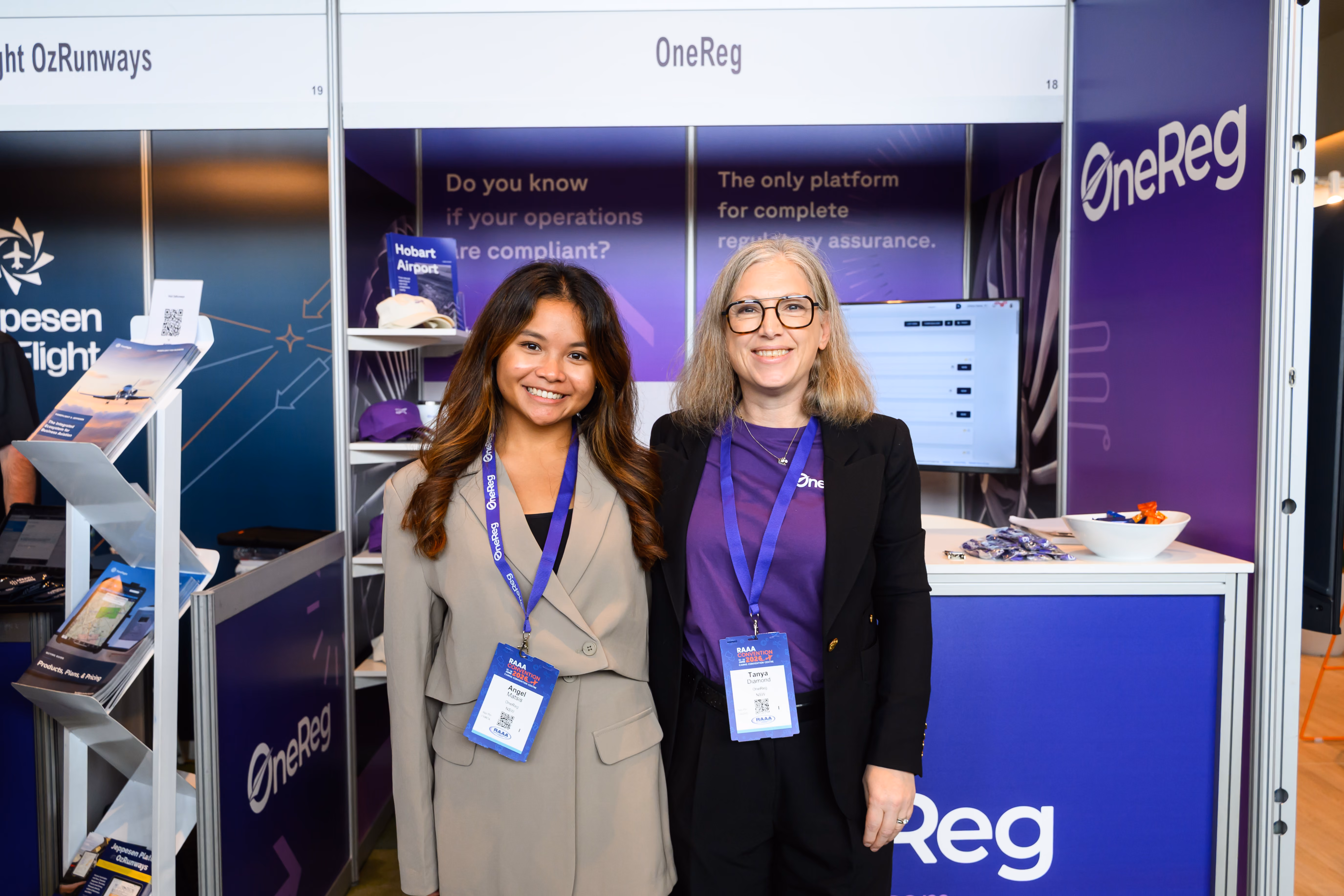 Two women with OneReg badges smiling at a purple OneReg booth at a convention or expo.