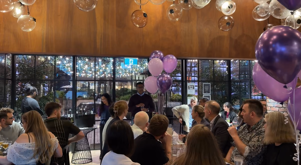 Group of people seated indoors around tables listening to a speaker with purple and pink balloons nearby.