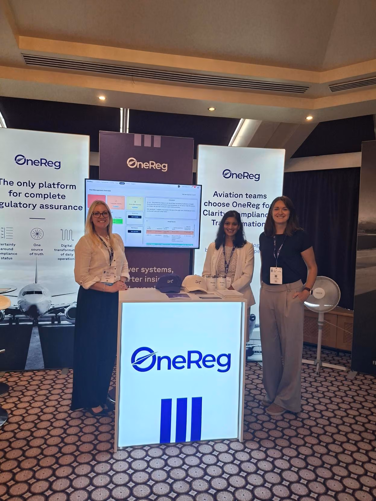 Three women standing at OneReg exhibition booth with aviation regulatory assurance displays.