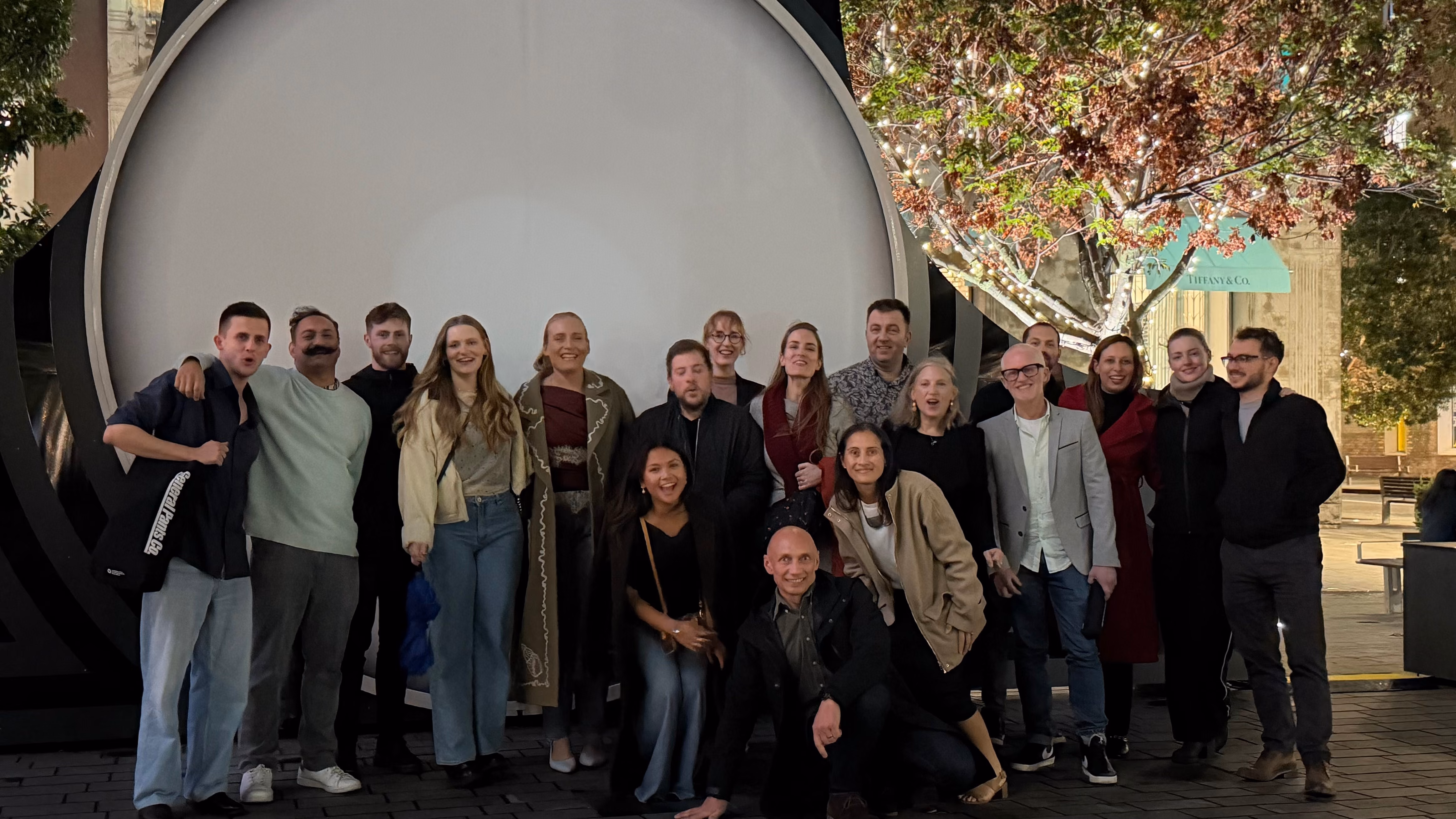 Group of 17 people posing and smiling outdoors in front of a large circular lighted backdrop at night.
