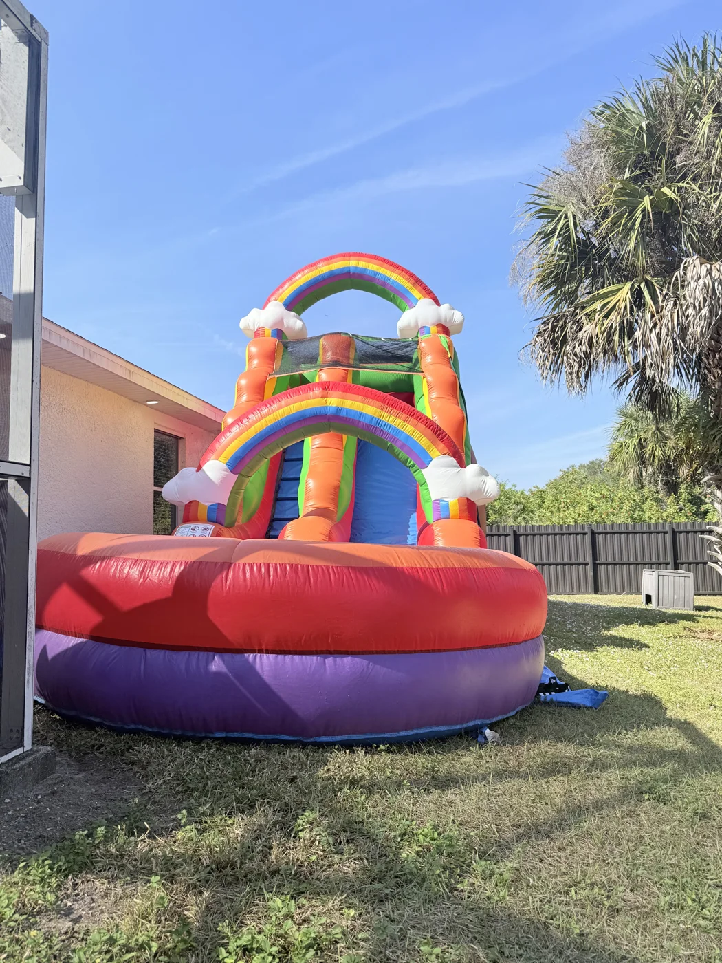 Rainbow-arched inflatable slide with cloud shapes and a broad red-and-purple basin on grass.