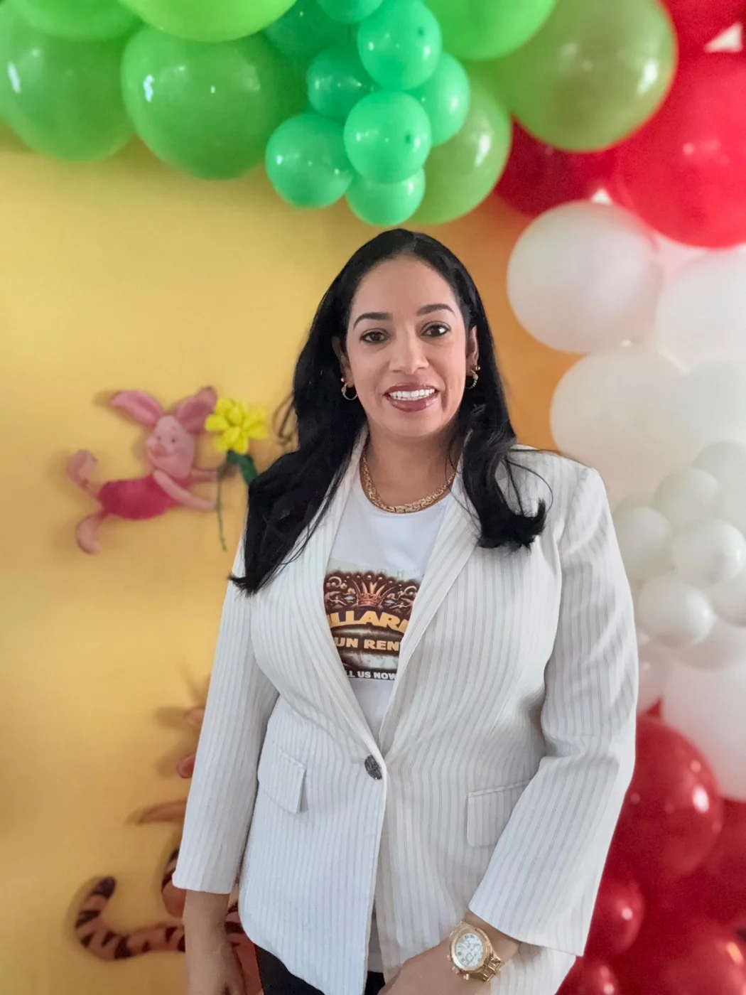 Smiling person in a light blazer in front of green, white and red balloons and character decorations.