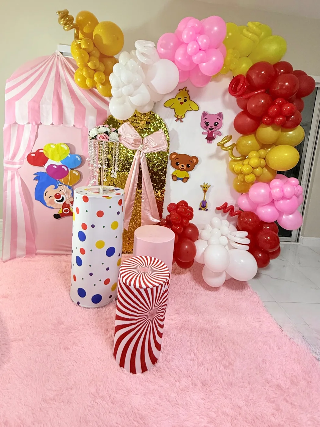Pink circus-themed display with striped panel, cartoon decorations, colorful balloon clusters and cylindrical stands.