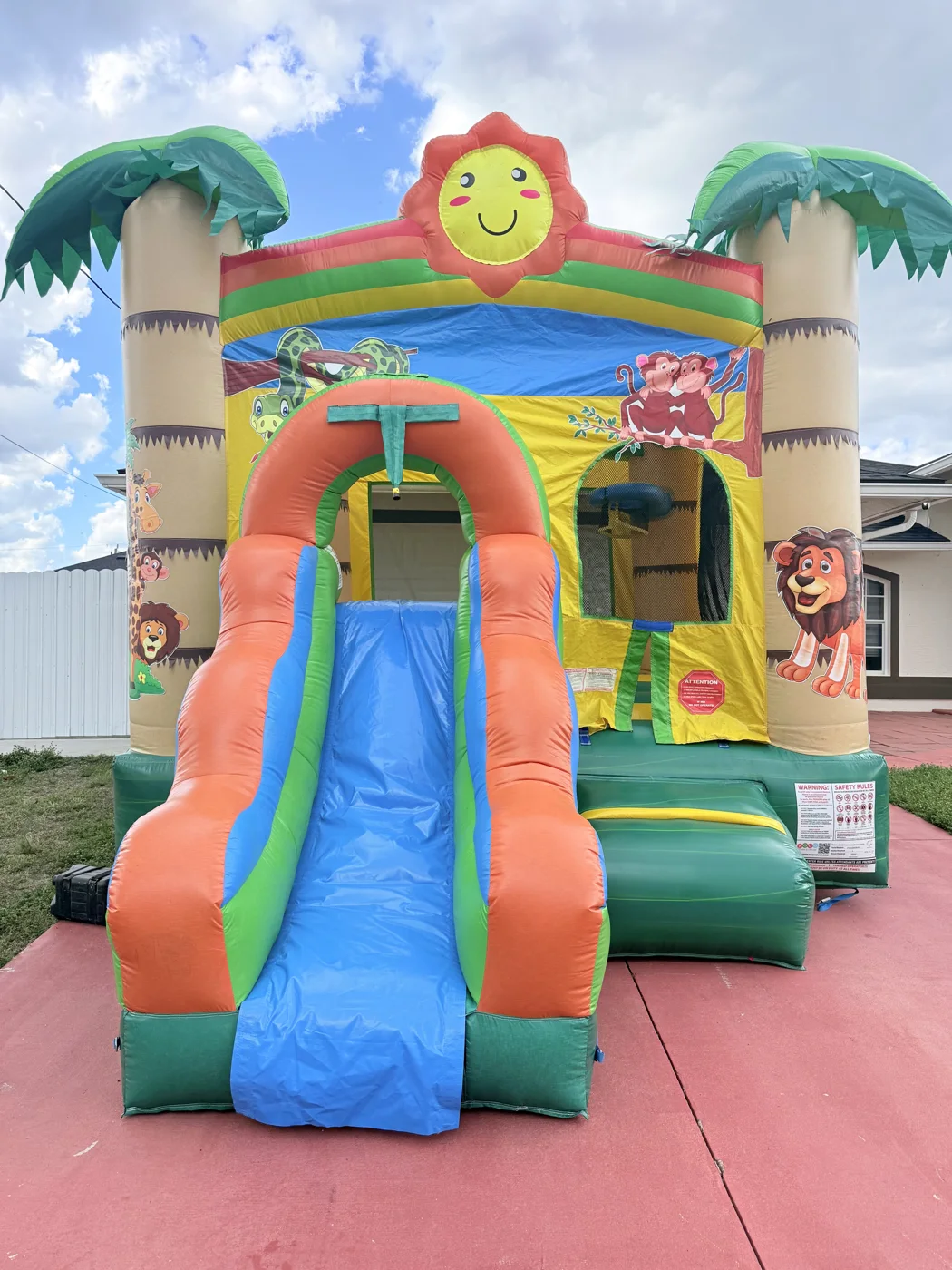 Tropical-themed inflatable with palm-shaped columns, cartoon animals, blue slide and green entrance.