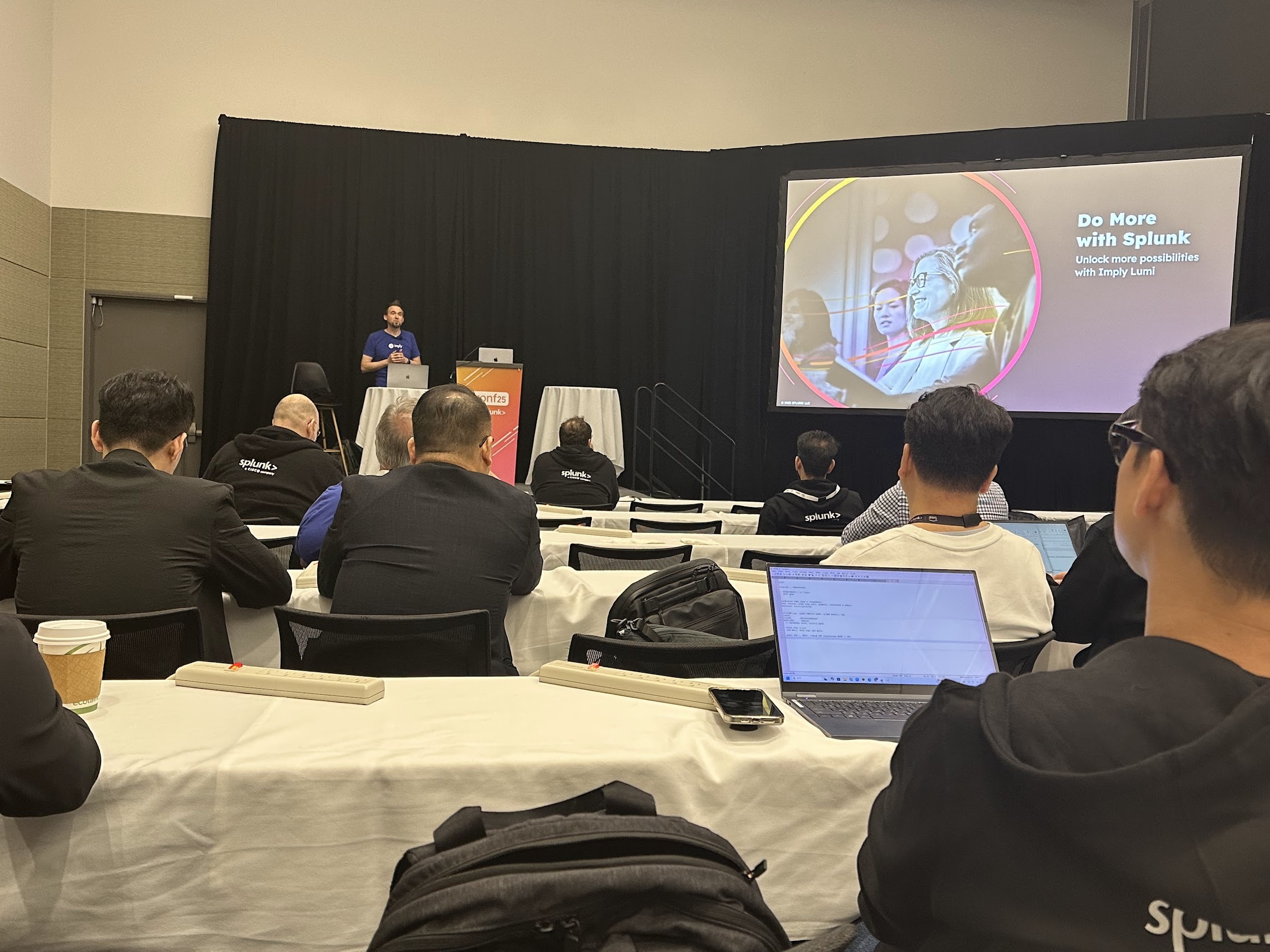 A speaker presenting a “Do more with Splunk” session on a projector screen to a seated audience at Splunk .conf25.
