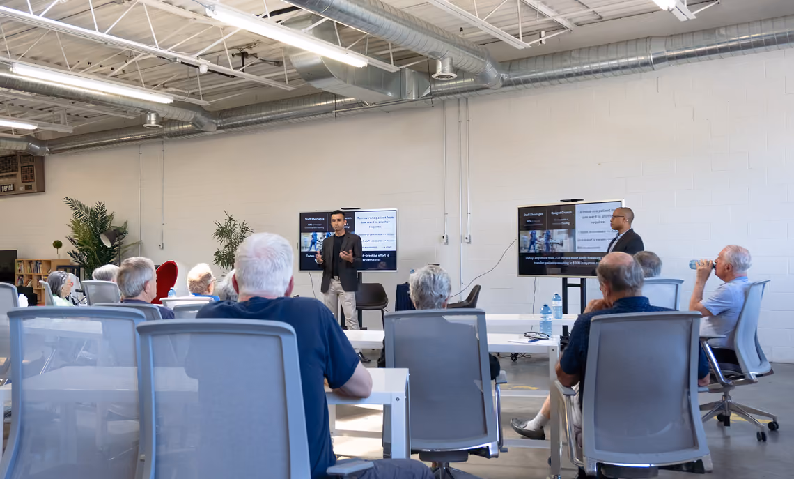 A group of people seated in an office or classroom setting facing two presenters standing near wall-mounted screens displaying presentation slides.